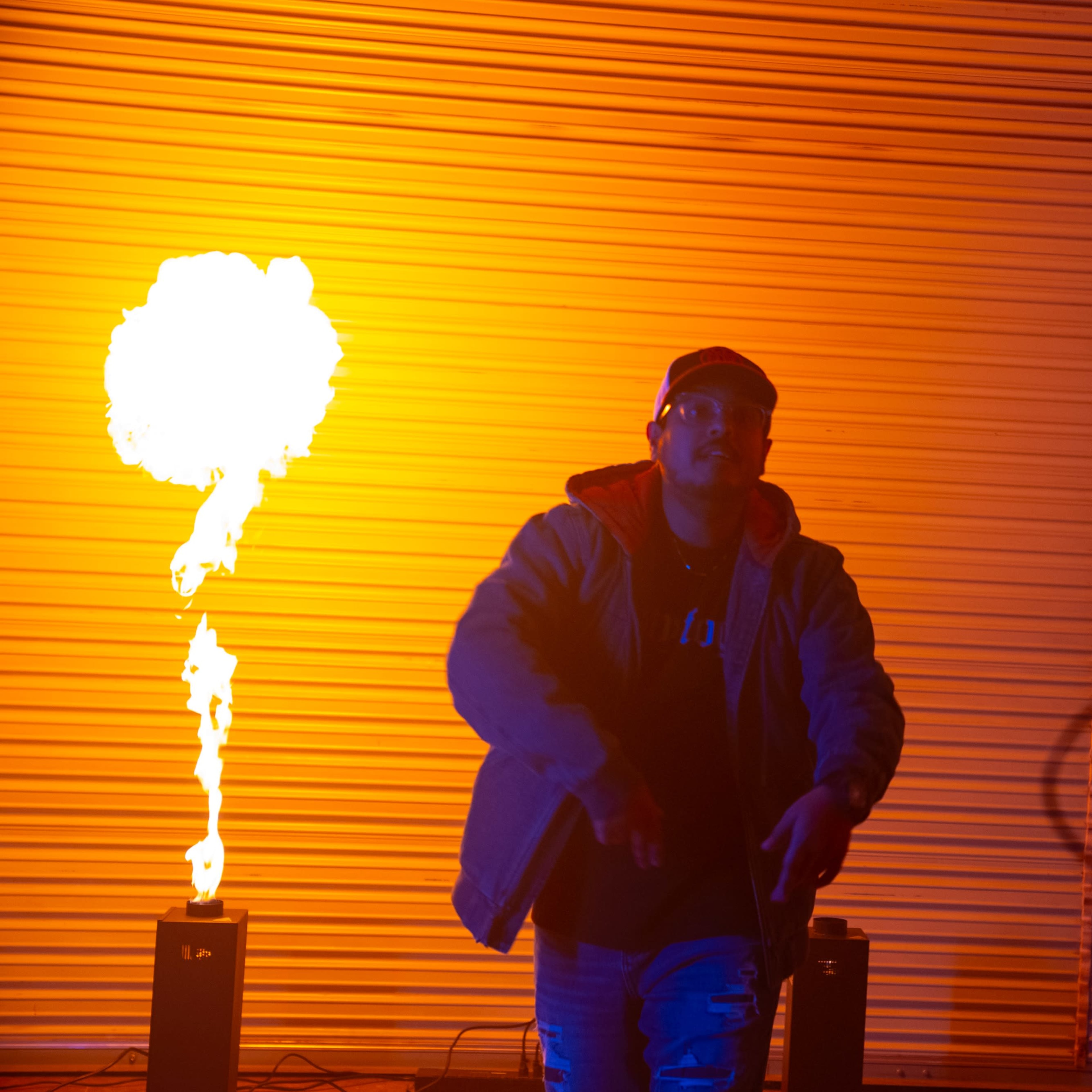 A rapper performs in front of a flame effect against a backdrop of a corrugated metal wall.
