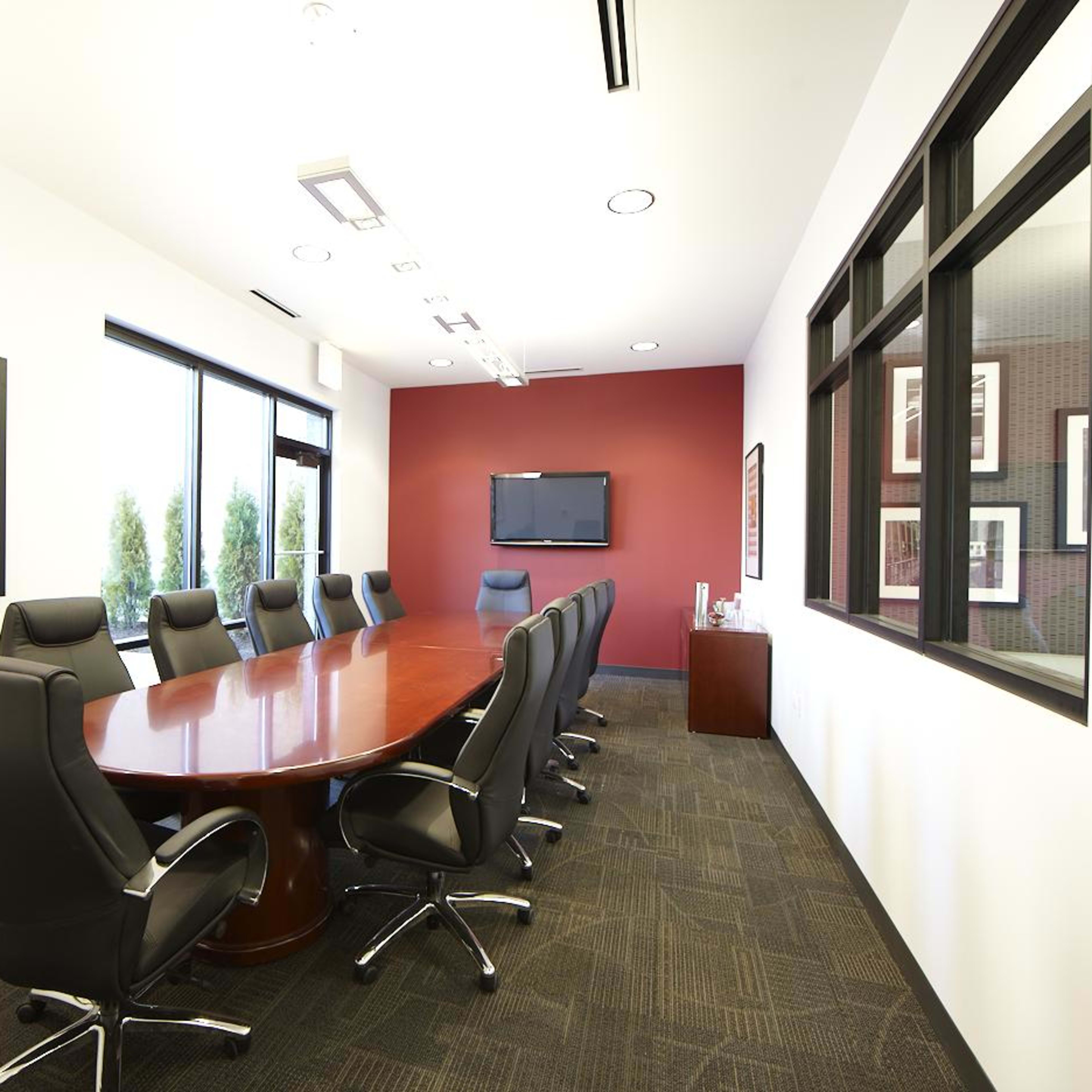 A spacious conference room features a large wooden table surrounded by black leather chairs and is equipped with a wall-mounted television and artwork.