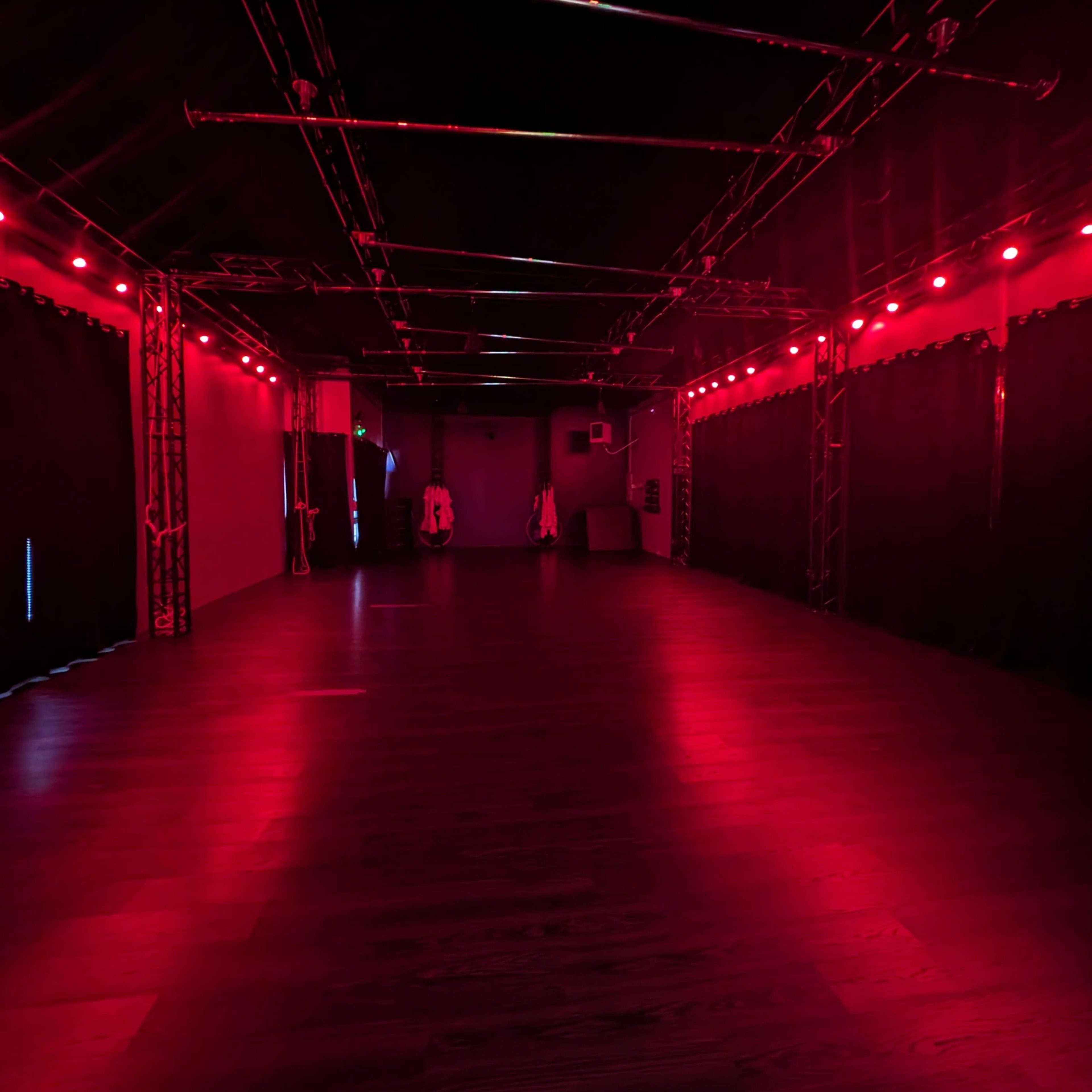 An empty room with a dark floor and red lighting, featuring black curtains and metal beams along the ceiling.