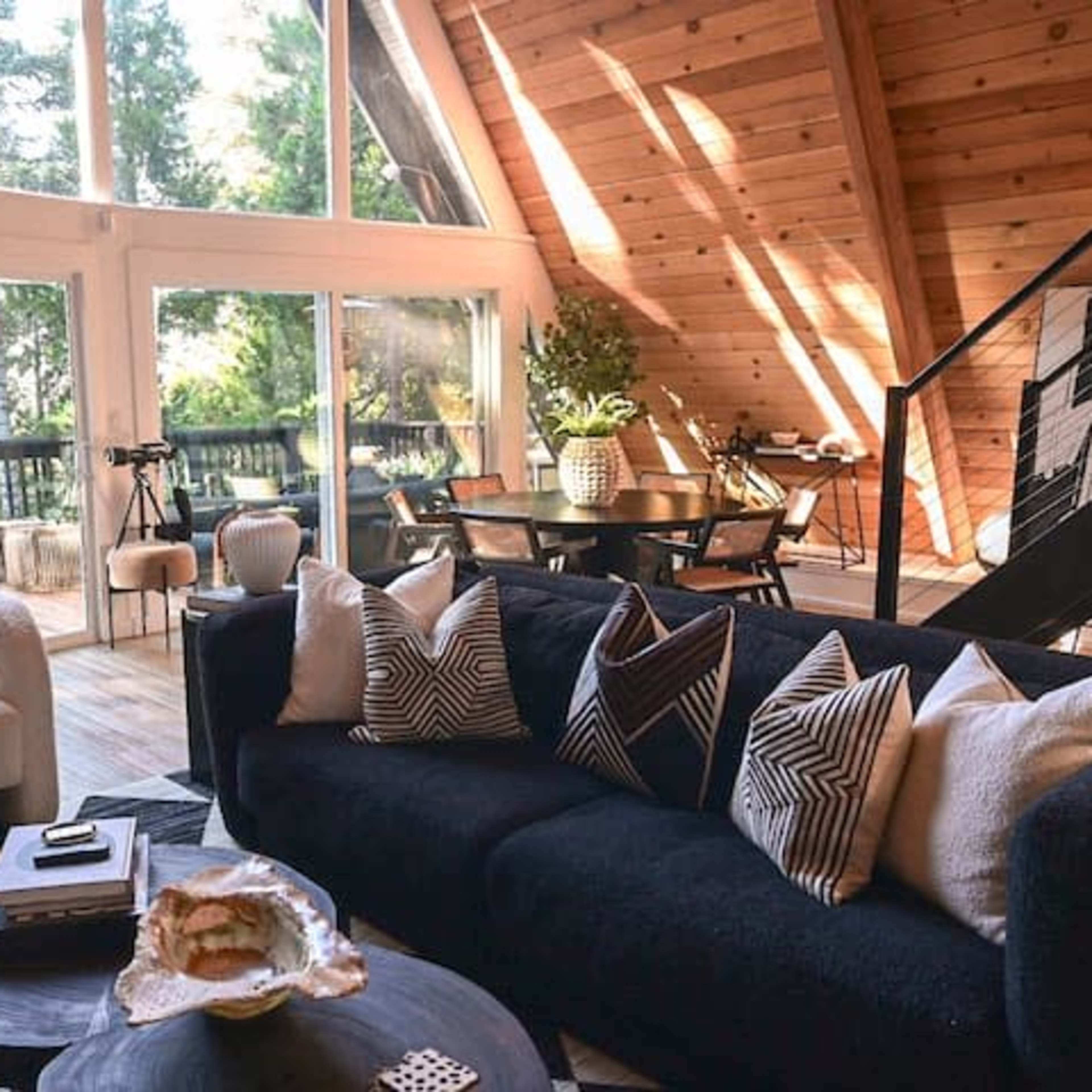 A modern living space with large windows, wooden walls, and a black sofa adorned with decorative pillows, surrounded by a dining area and greenery.