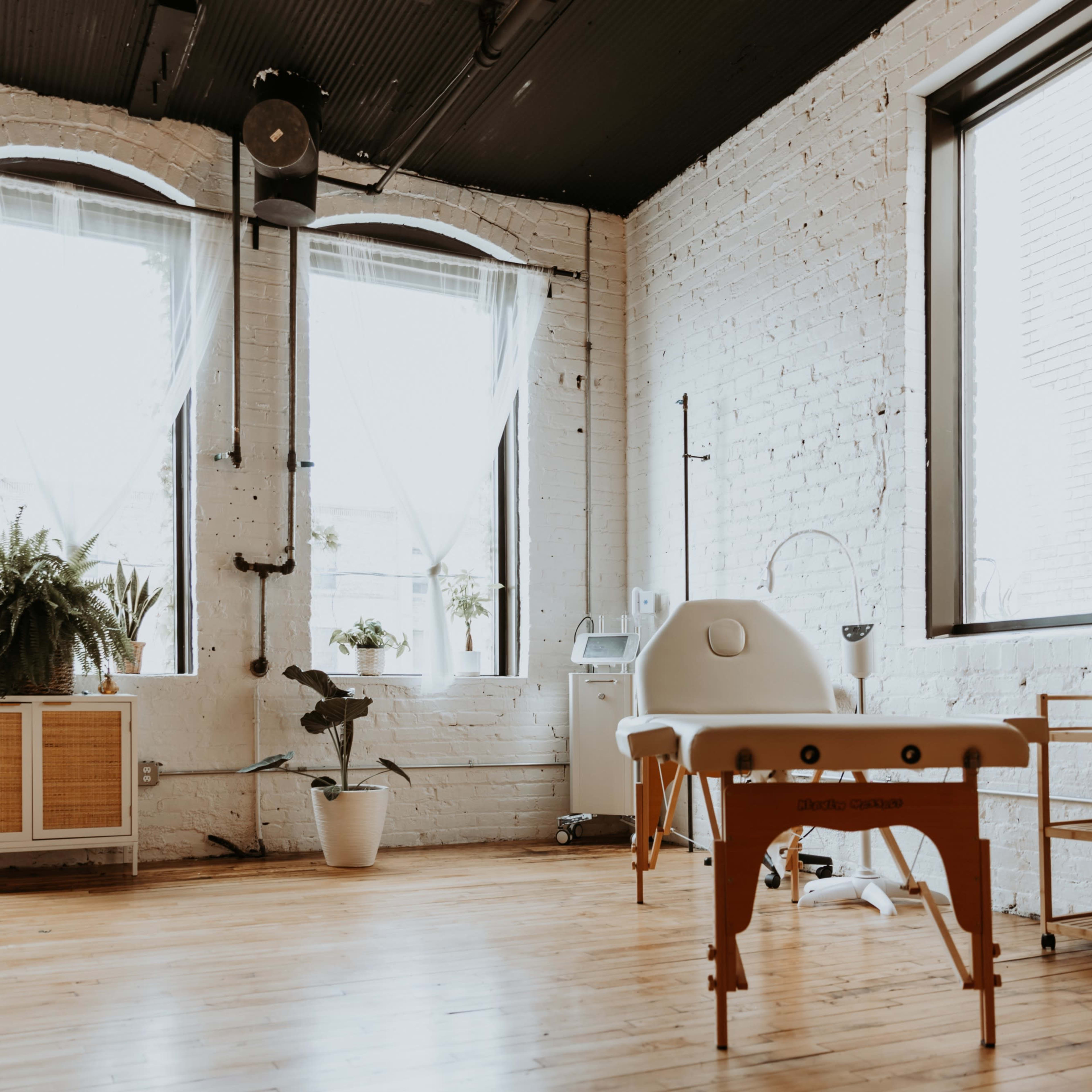 A bright, minimalistic treatment room featuring a massage table, large windows with sheer curtains, and plants arranged artistically.