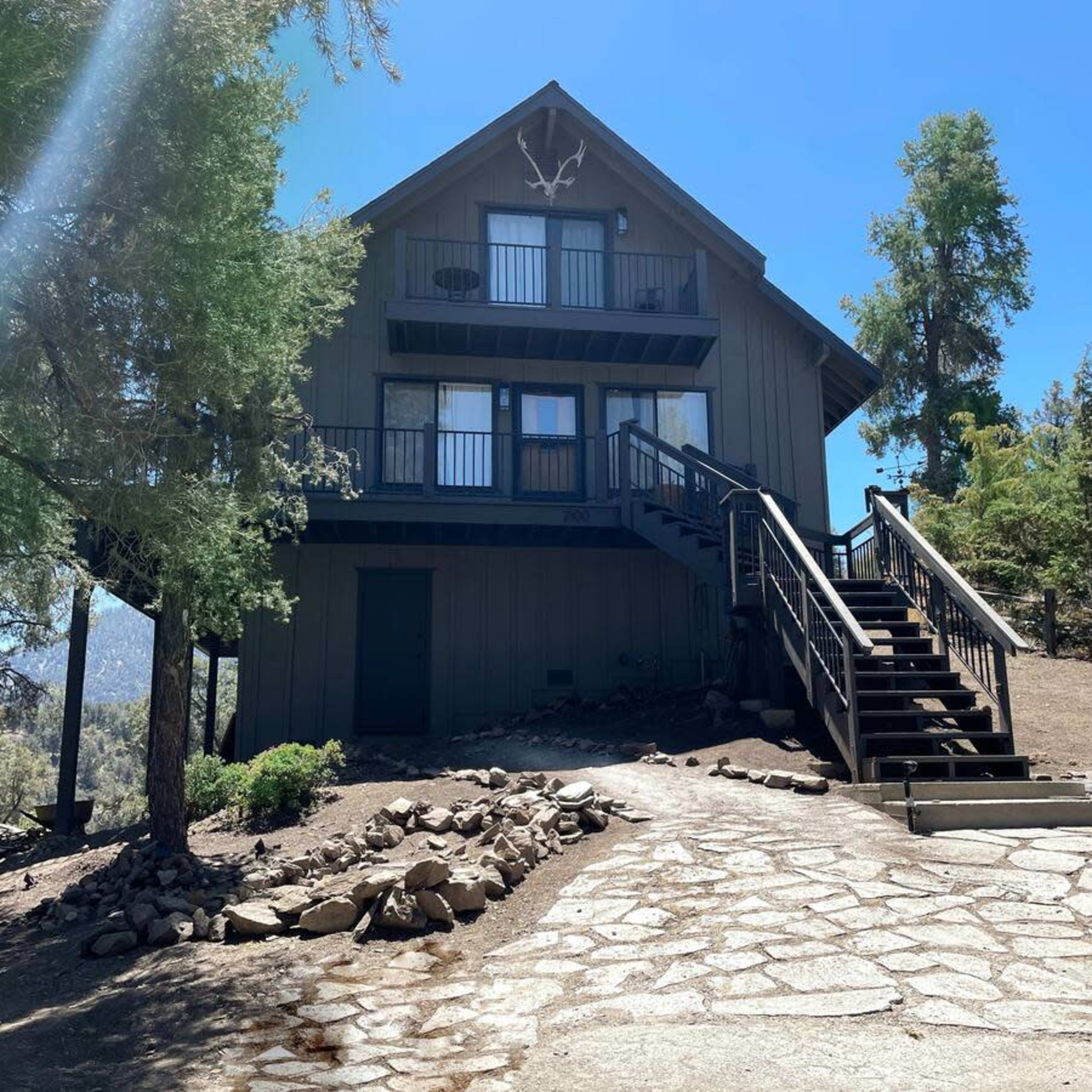 A three-story cabin with a wooden exterior and a set of stairs leads up to a covered porch surrounded by trees.