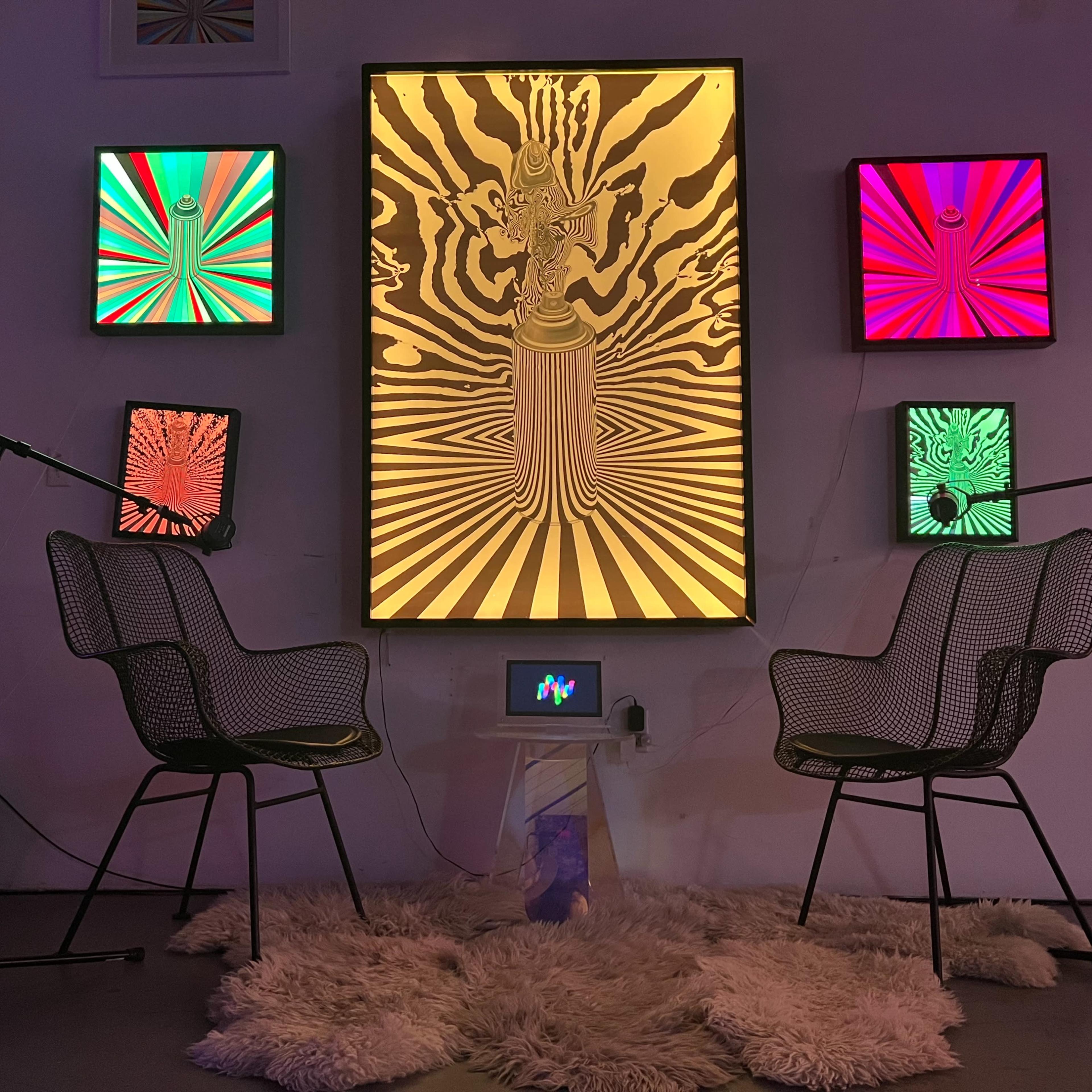 A colorful art installation featuring a central illuminated piece with a design of a figure, flanked by smaller vibrant screens, with two wire chairs facing a small display on a rug.