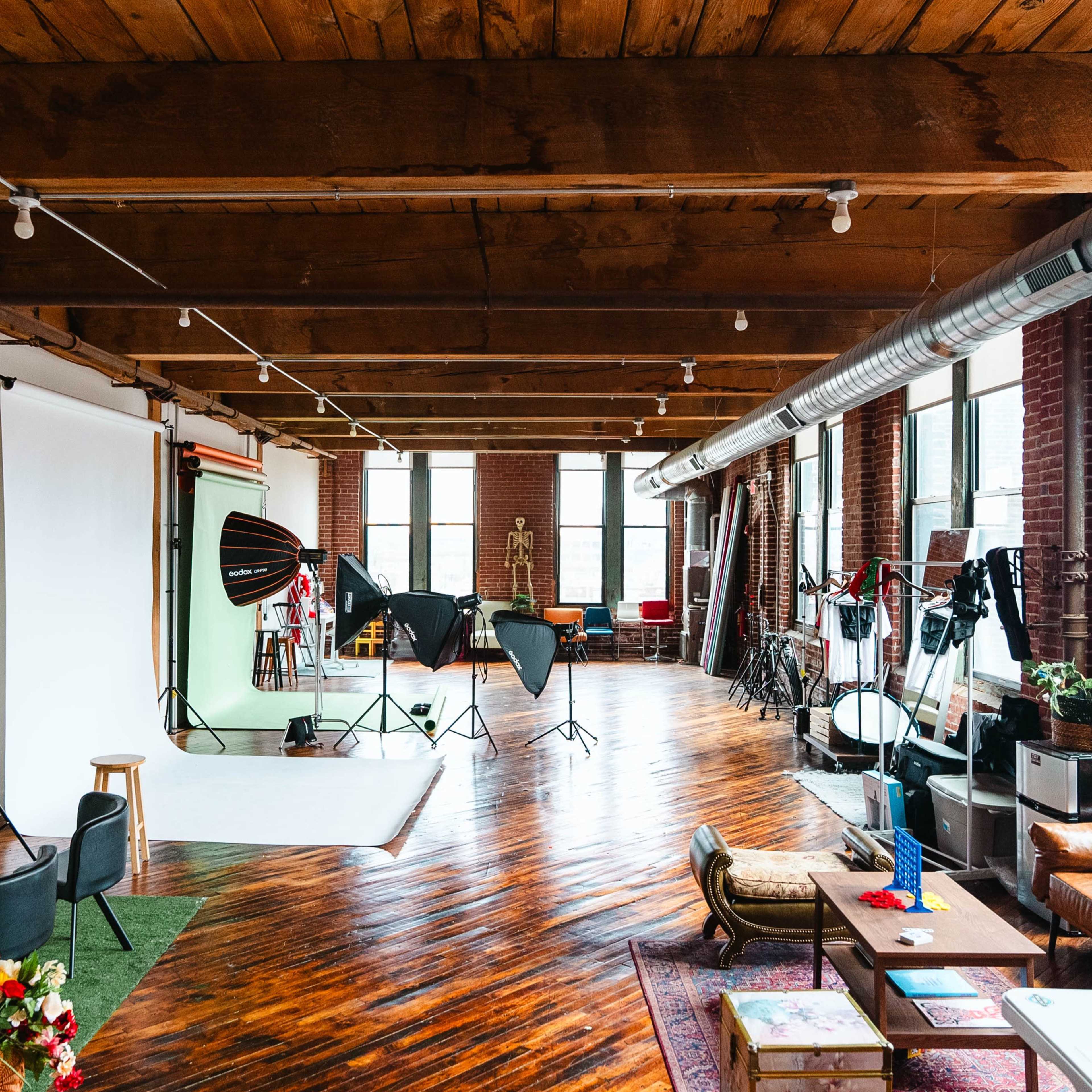 A spacious photo studio with wooden floors, large windows, and various lighting equipment set up around a backdrop.