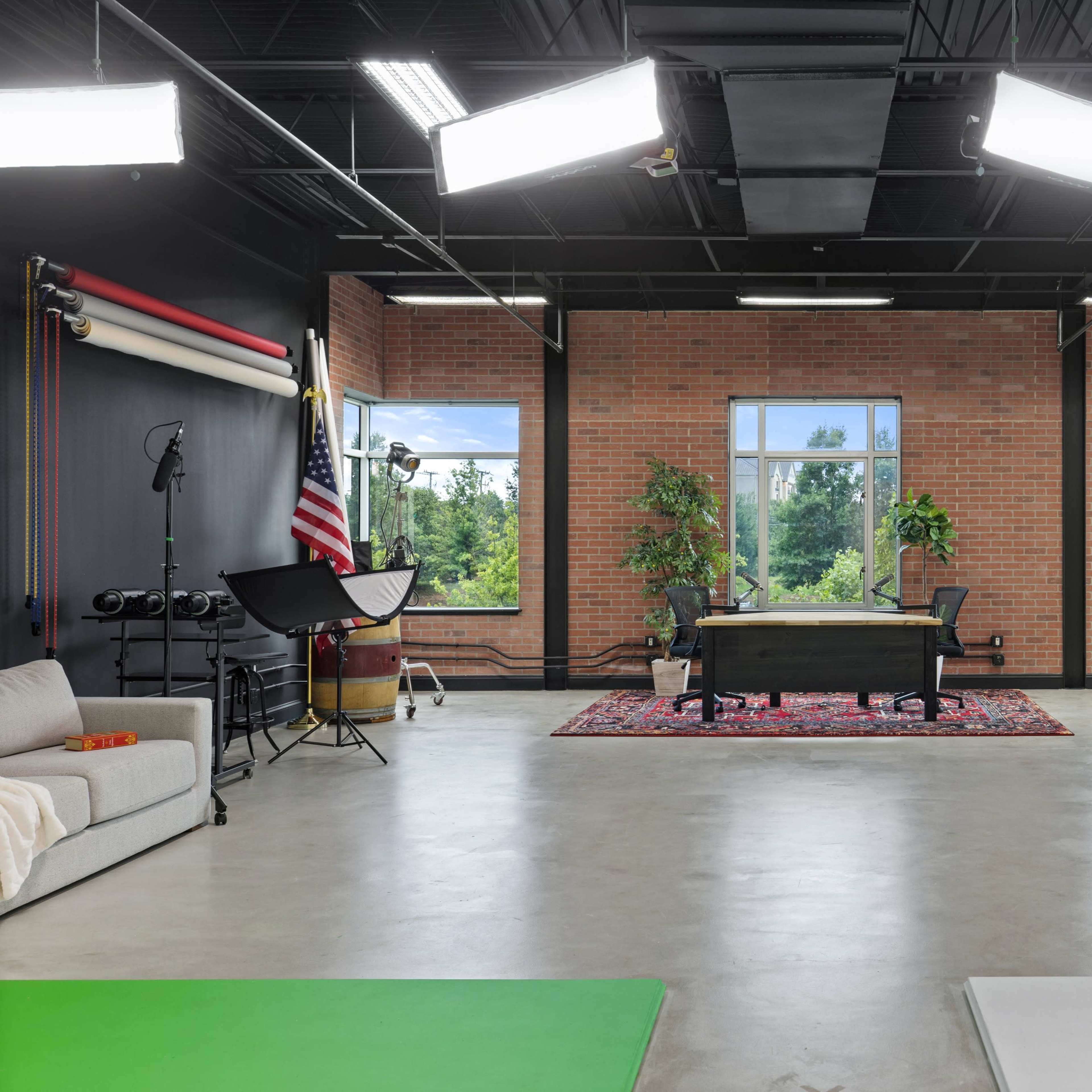A spacious studio with large windows, a wooden desk at the center, seating areas, and various lighting equipment.