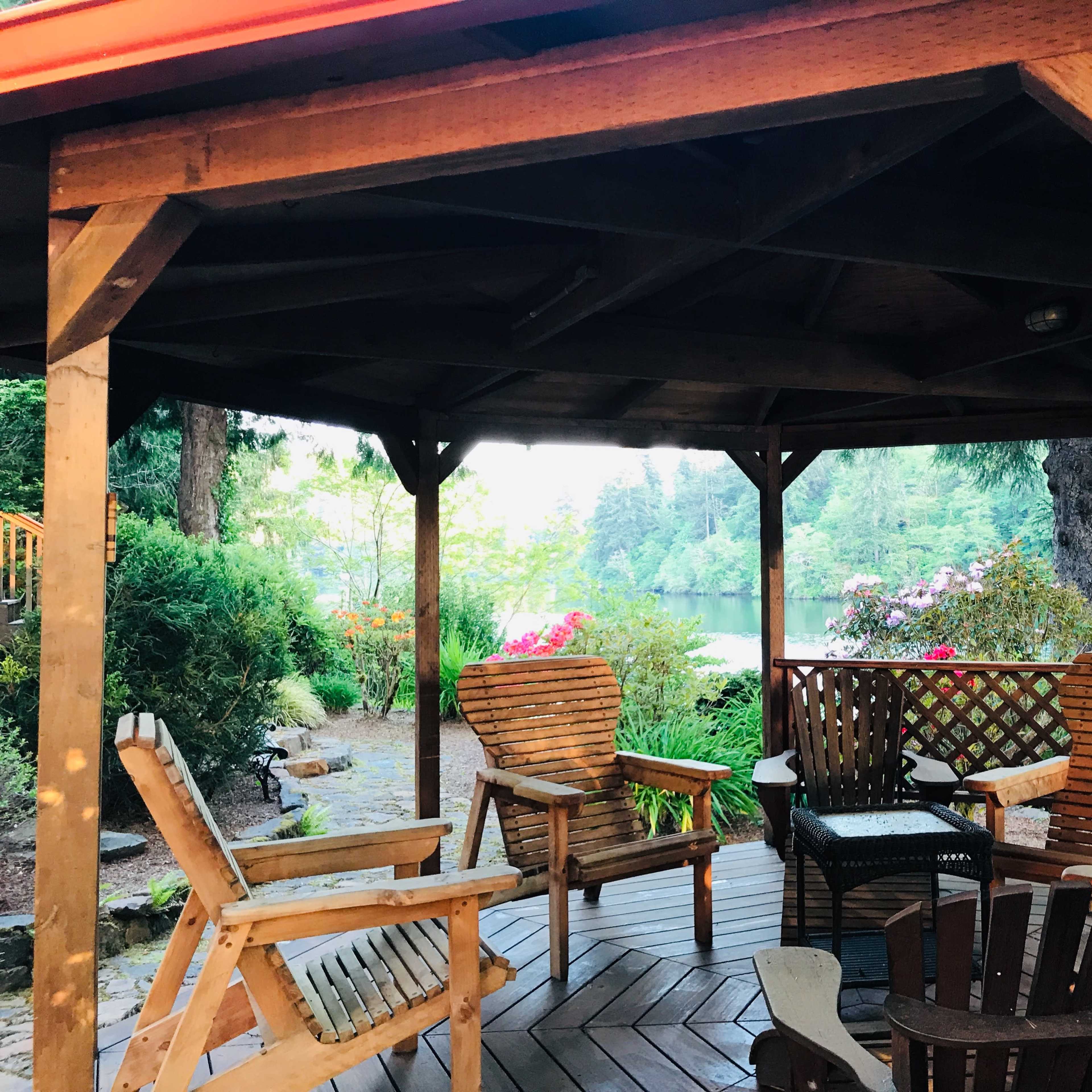 A wooden gazebo with several chairs overlooks a calm lake surrounded by trees and blooming flowers.