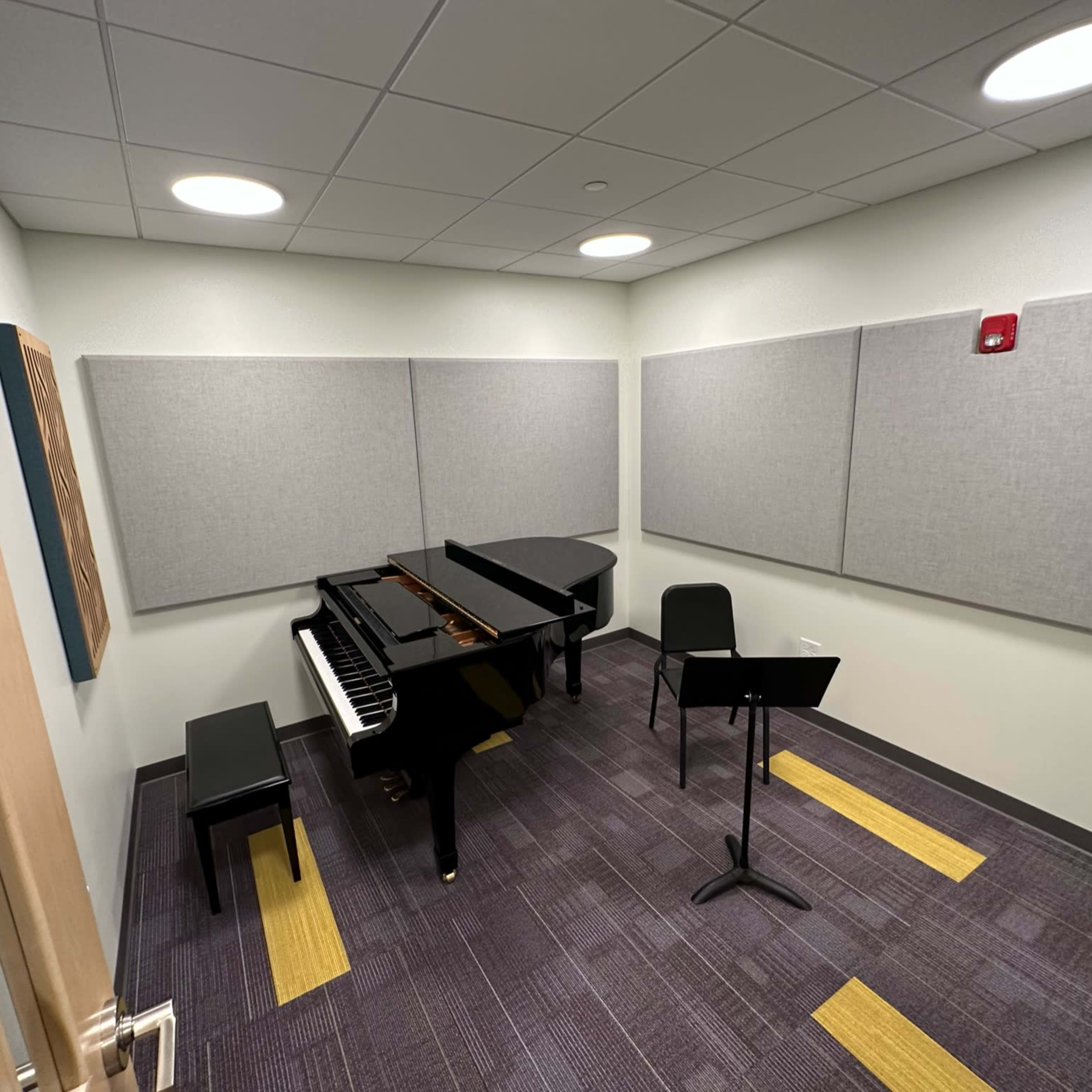 A small music practice room featuring a grand piano, a chair, and a music stand, with sound-absorbing panels on the walls.