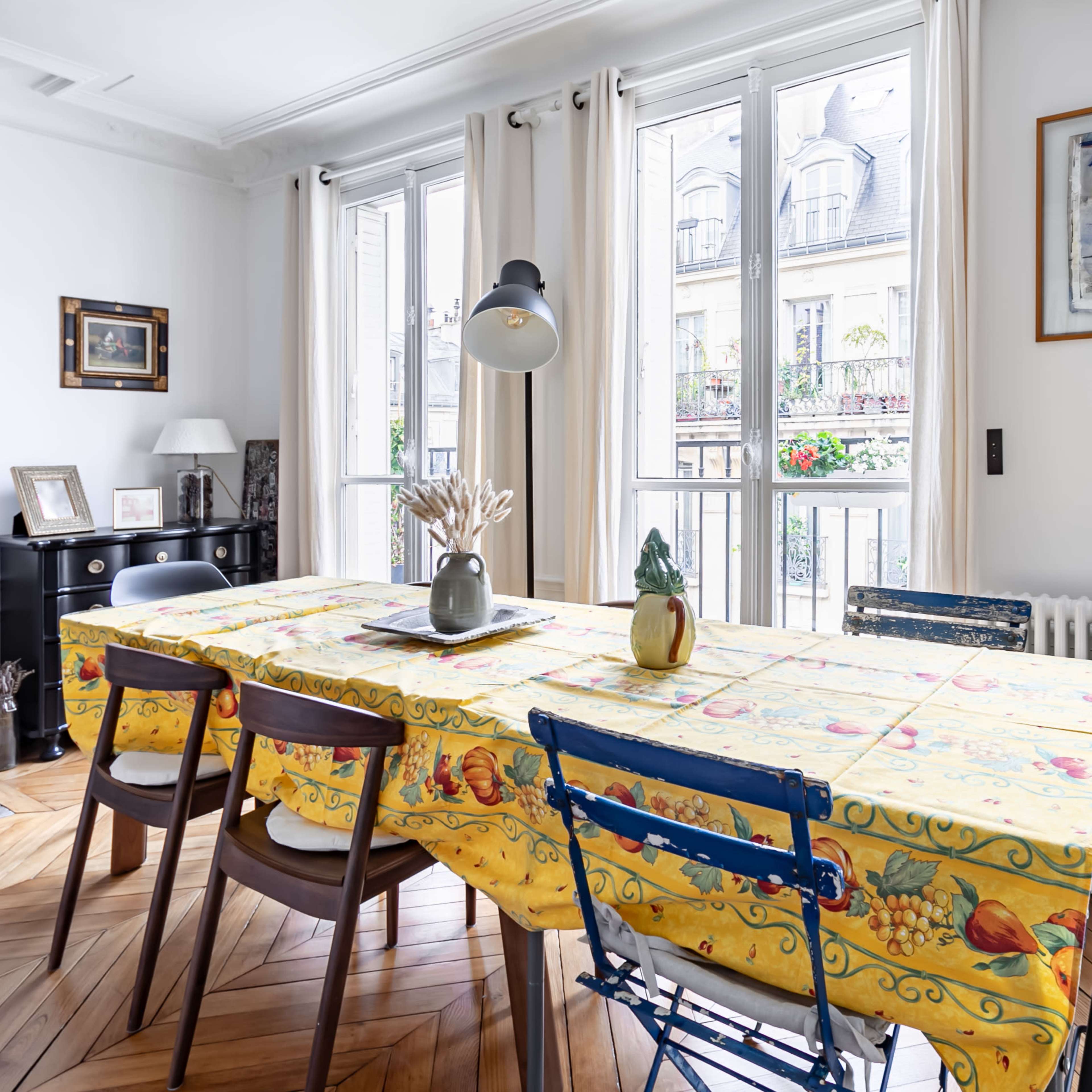 A dining room features a large table covered with a yellow tablecloth, surrounded by chairs, and illuminated by natural light from large windows.