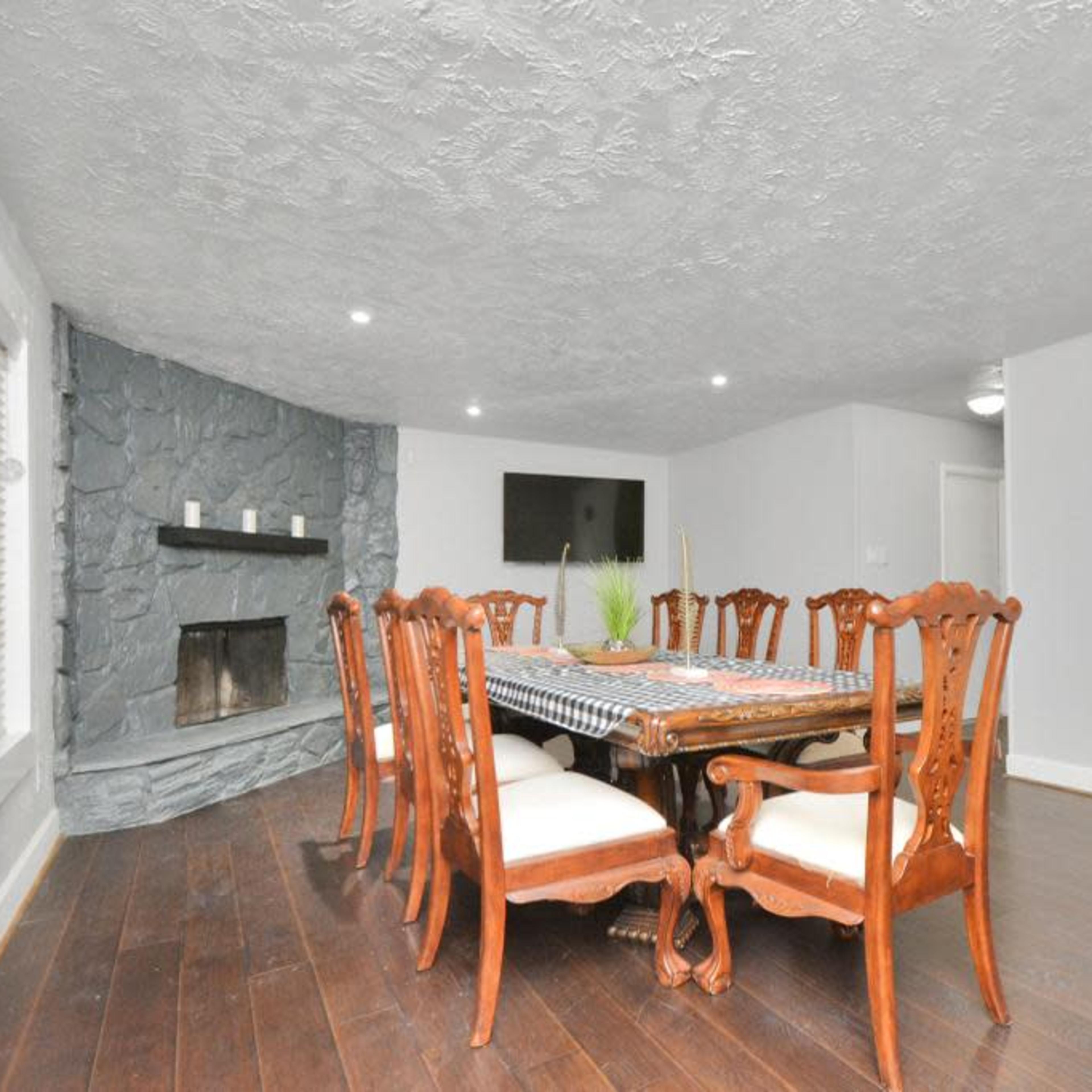 A dining room features a large wooden table surrounded by ornate chairs, with a stone fireplace and a TV mounted on the wall.