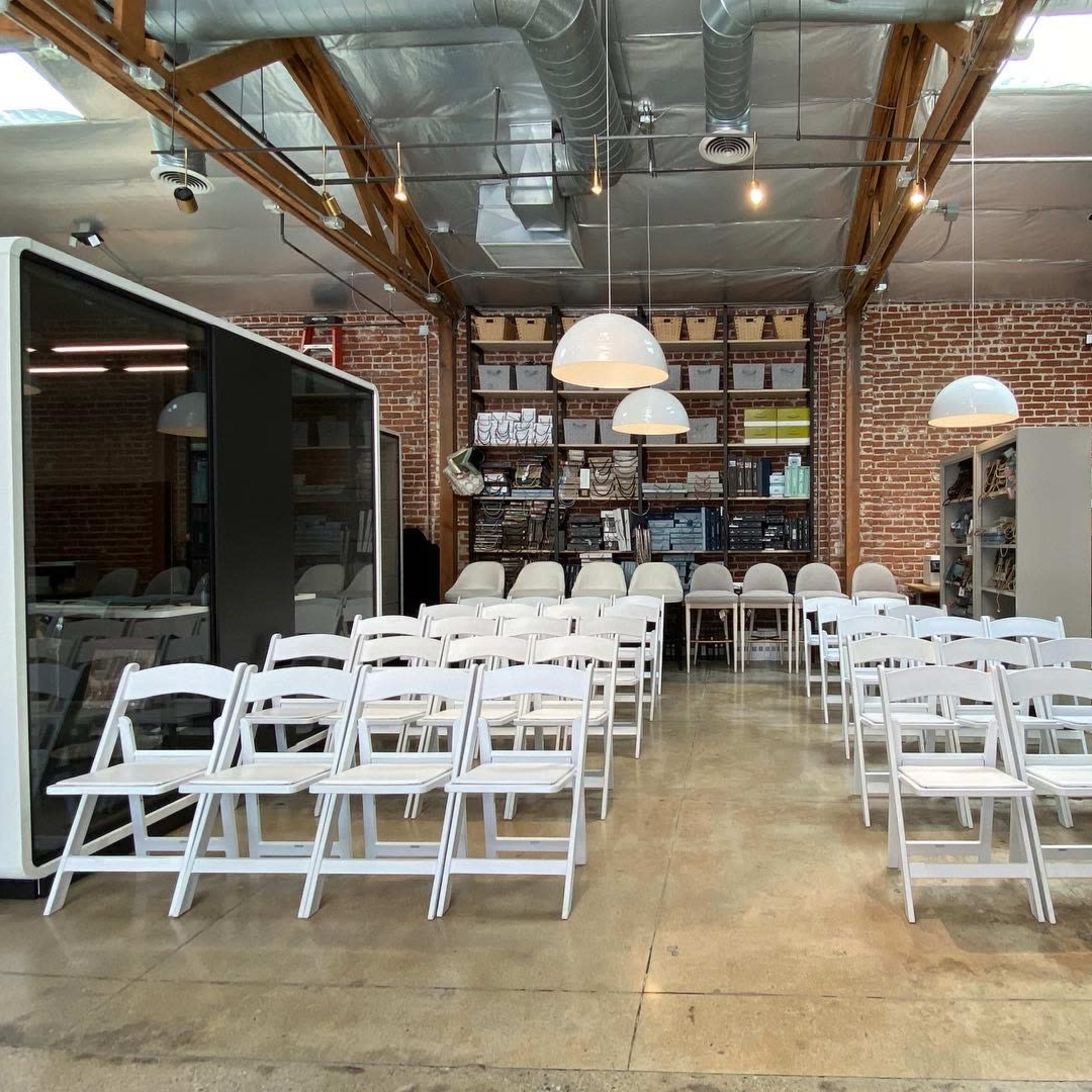 A spacious interior with rows of white folding chairs arranged for an event, against a backdrop of exposed brick walls and industrial shelving.