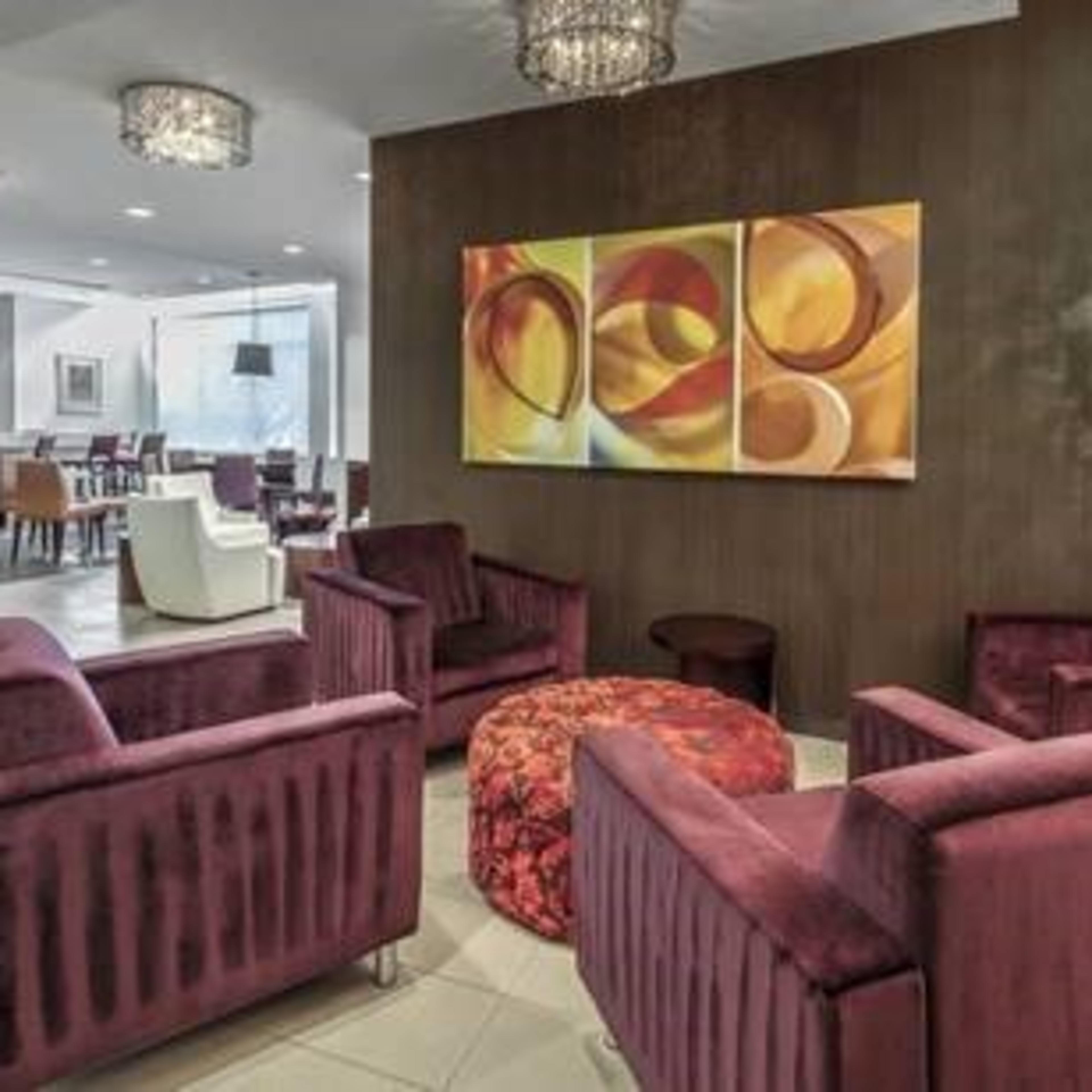 A modern lounge area featuring sleek purple armchairs, a round patterned ottoman, and decorative wall art under stylish light fixtures.