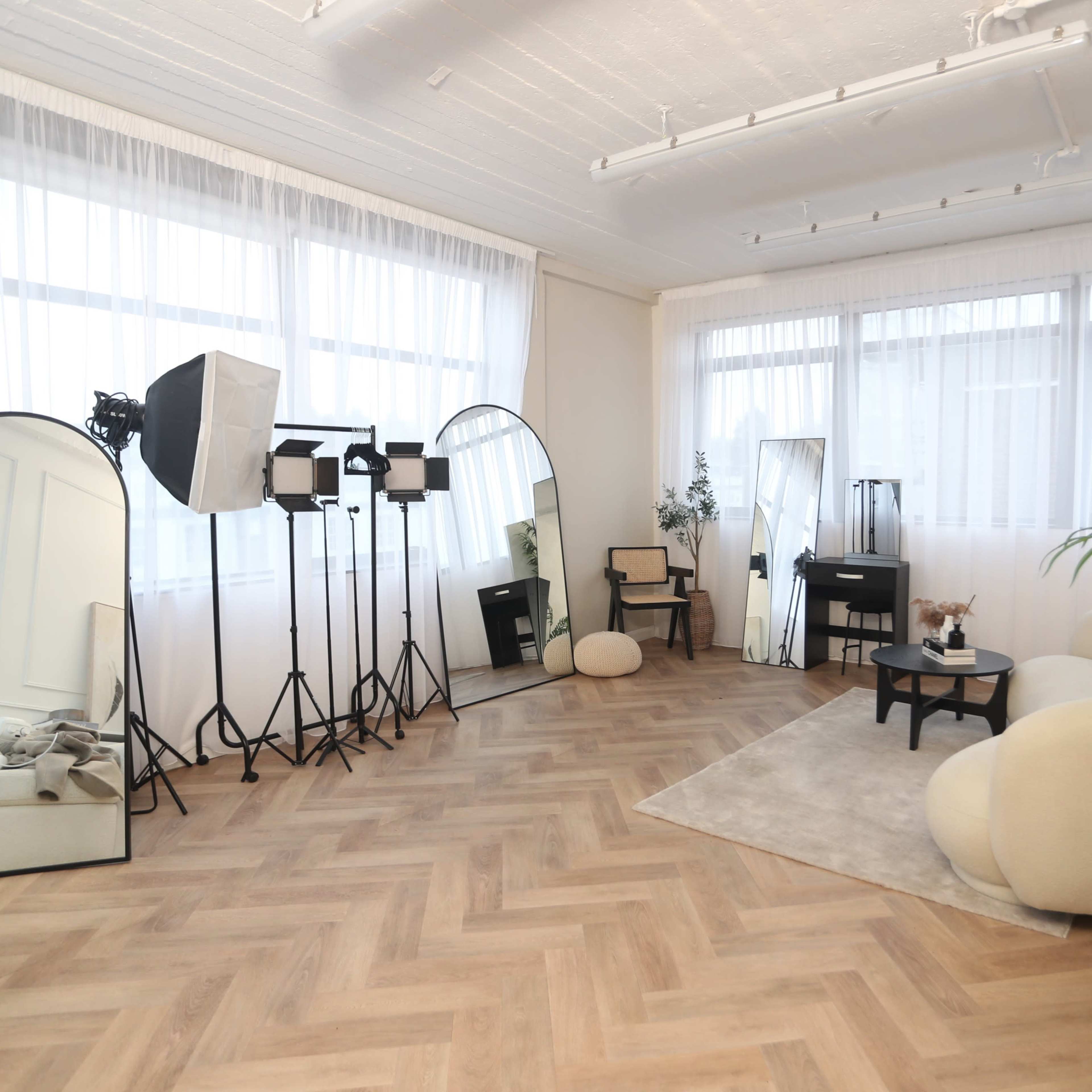 A bright studio space with large windows, featuring mirrors, soft furniture, and photography lighting equipment.