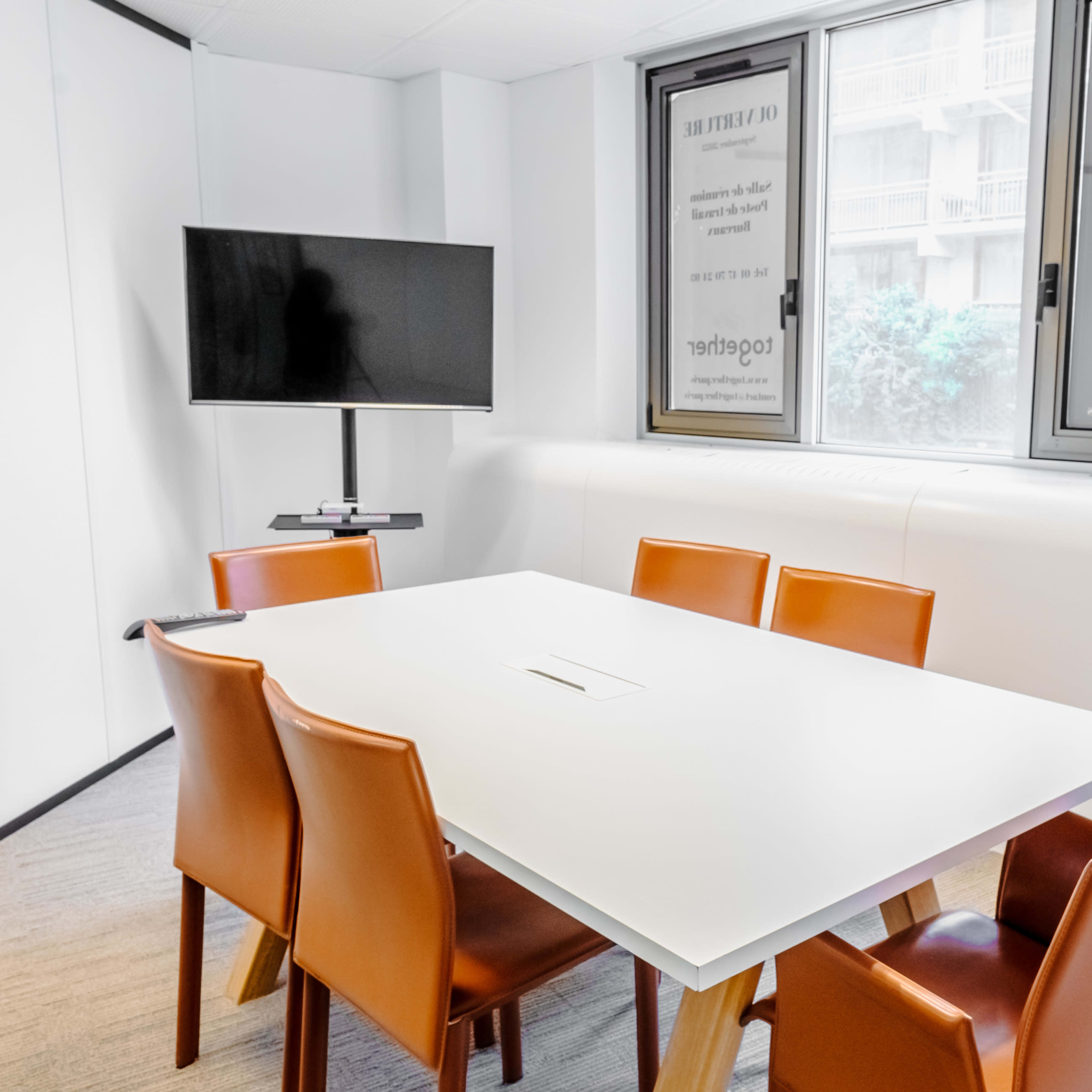 A modern conference room features a rectangular white table surrounded by six orange chairs, with a television mounted on the wall and large windows offering natural light.