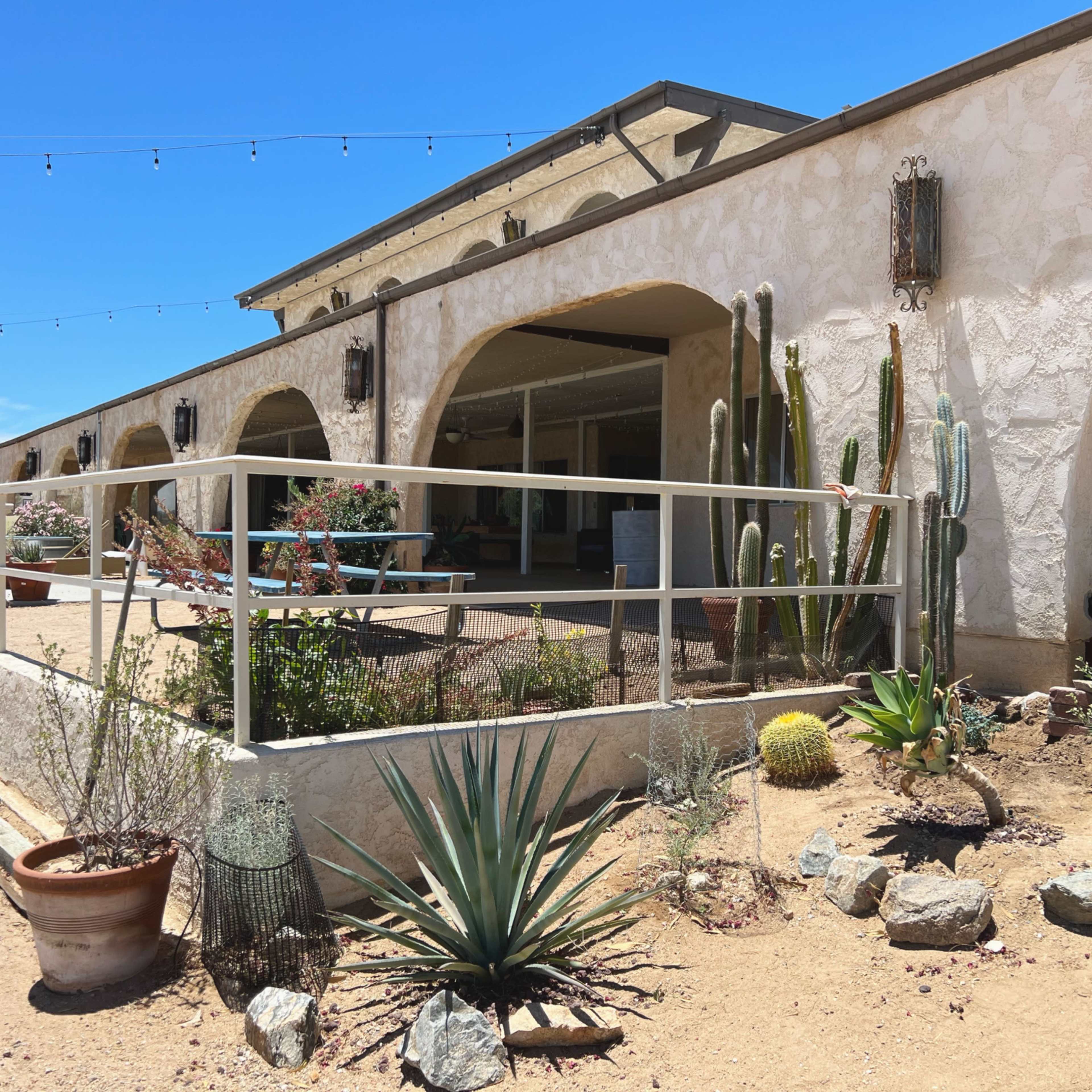 A single-story building with arched doorways and a landscaped area featuring cacti and various plants in a dry, sandy environment.