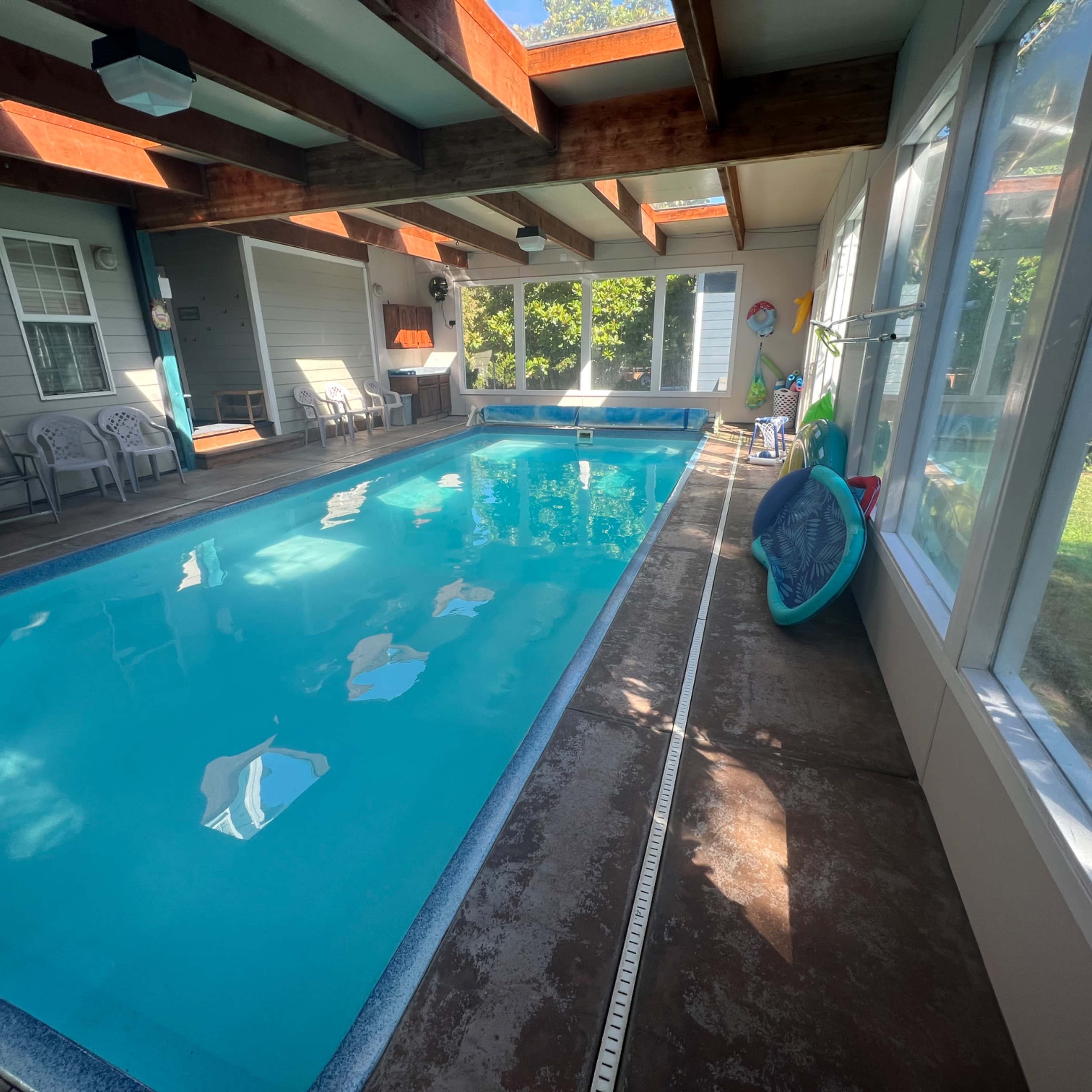 An indoor swimming pool surrounded by large windows and wooden beams, with a seating area beside it.