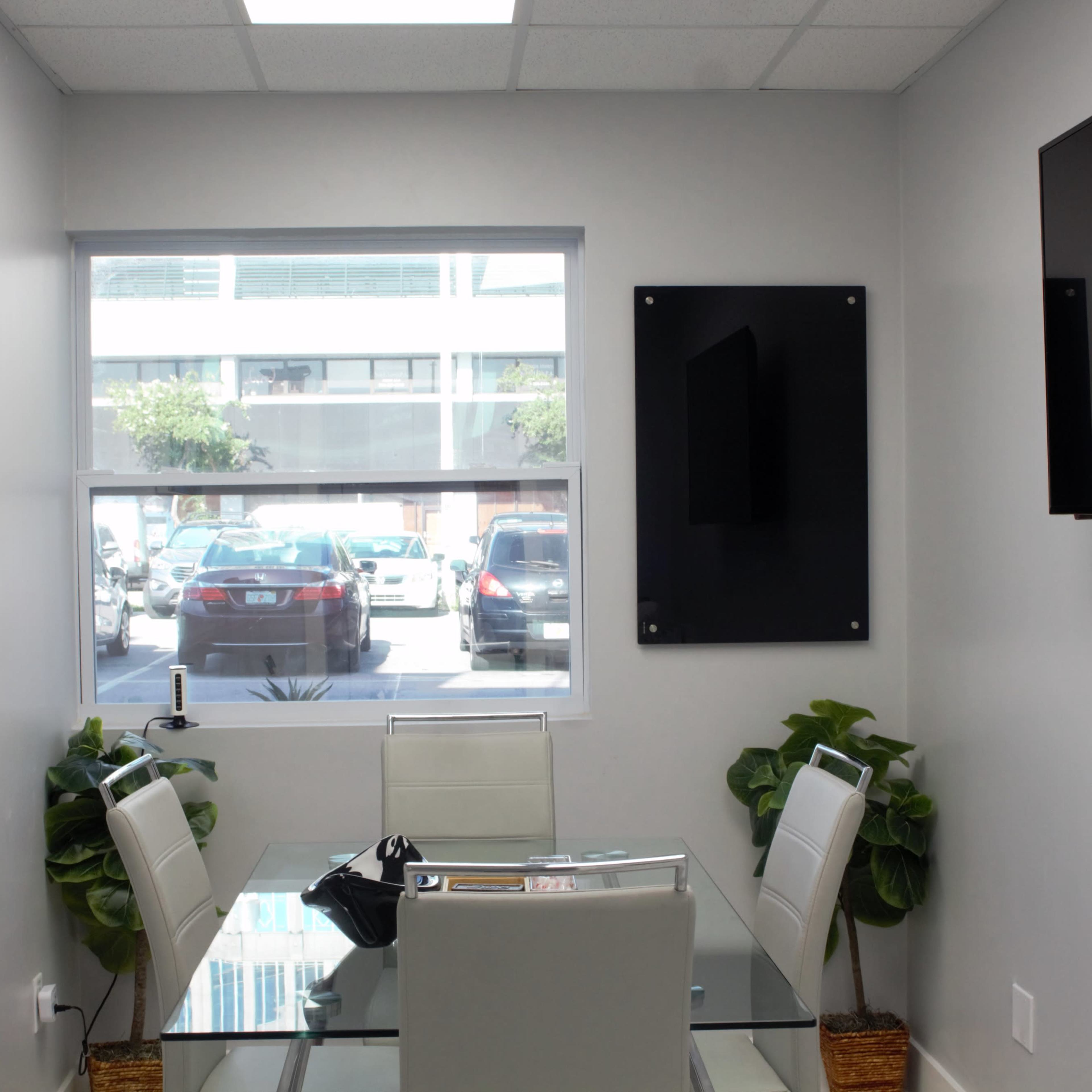 A brightly lit meeting room features a glass table with white chairs, surrounded by plants, and a view of parked cars outside.