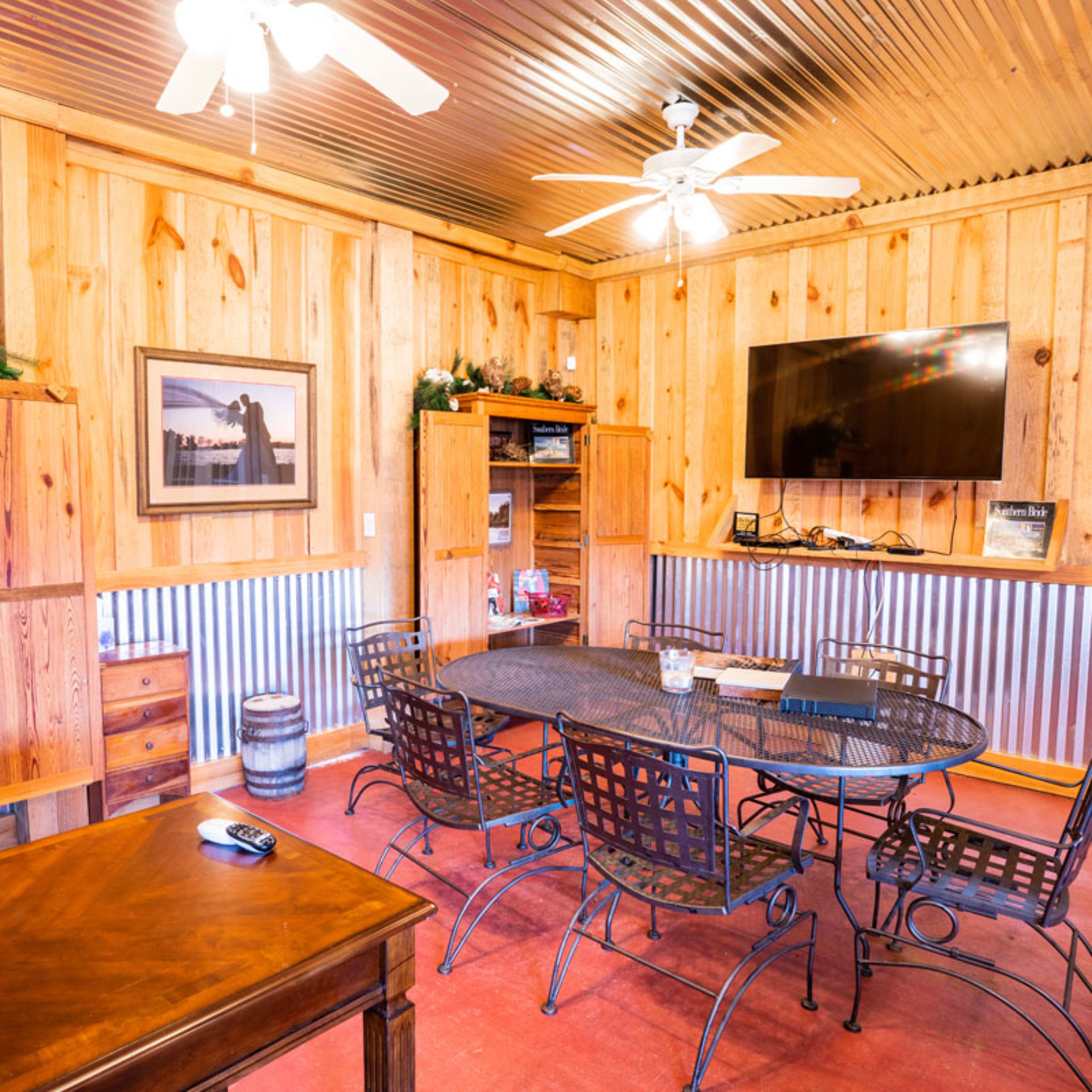 The interior of a wooden-paneled room features a round metal table surrounded by chairs, a TV mounted on the wall, and wooden shelving with decorative items.