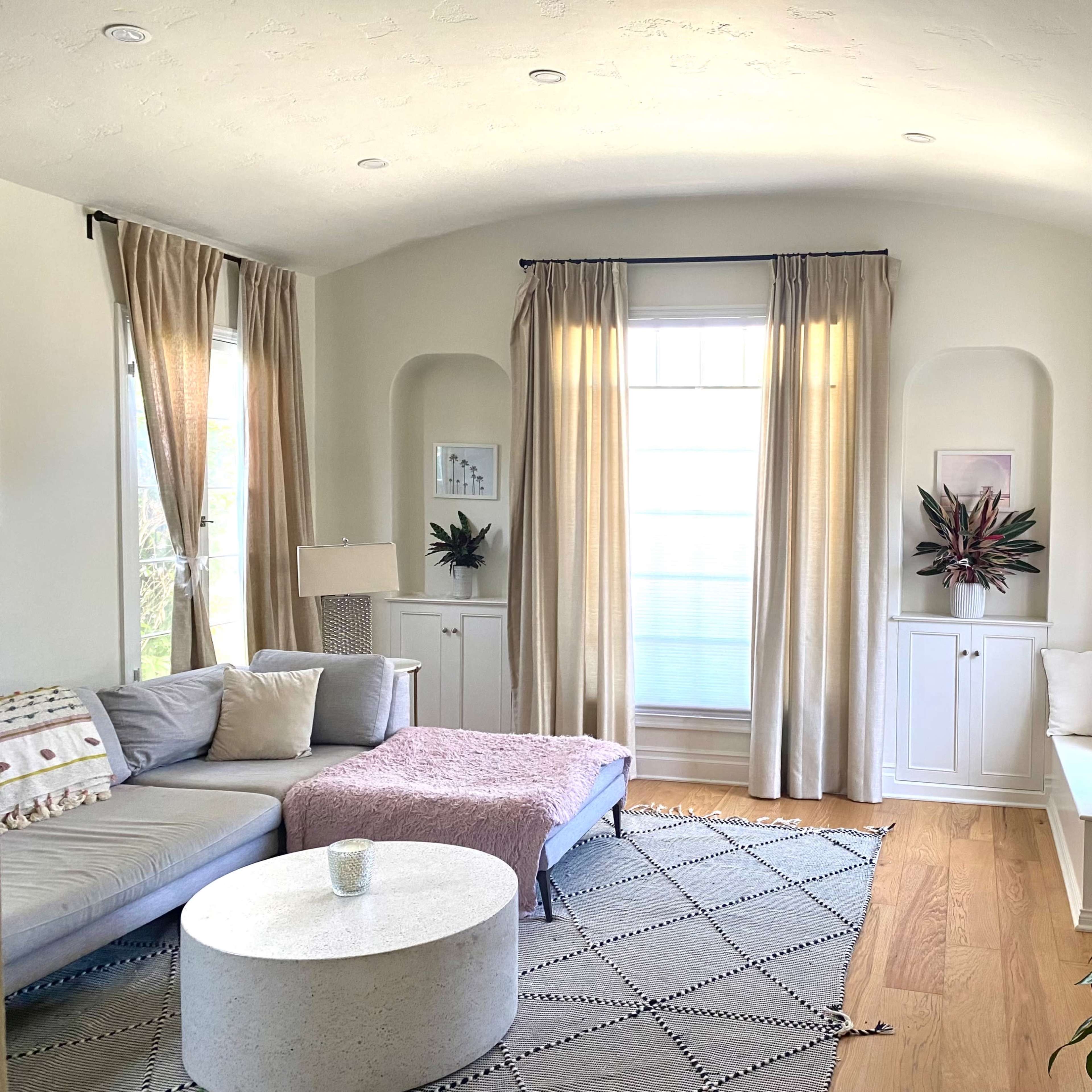 A bright living room with a curved ceiling, featuring a gray sectional sofa, a round coffee table, and a mounted television above a fireplace, complemented by large windows dressed with light curtains.