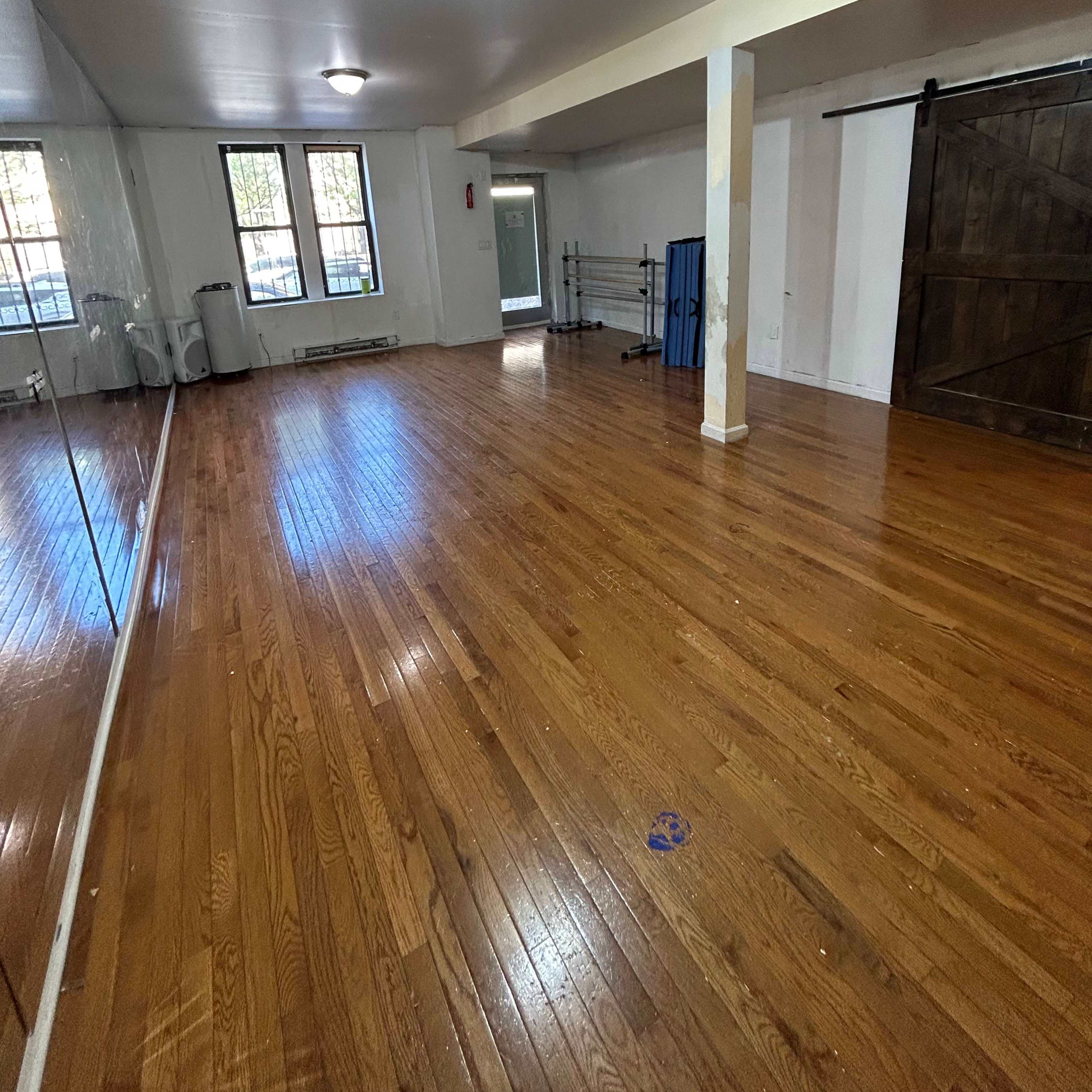 An empty room with hardwood flooring, large windows, and a mirrored wall on one side.
