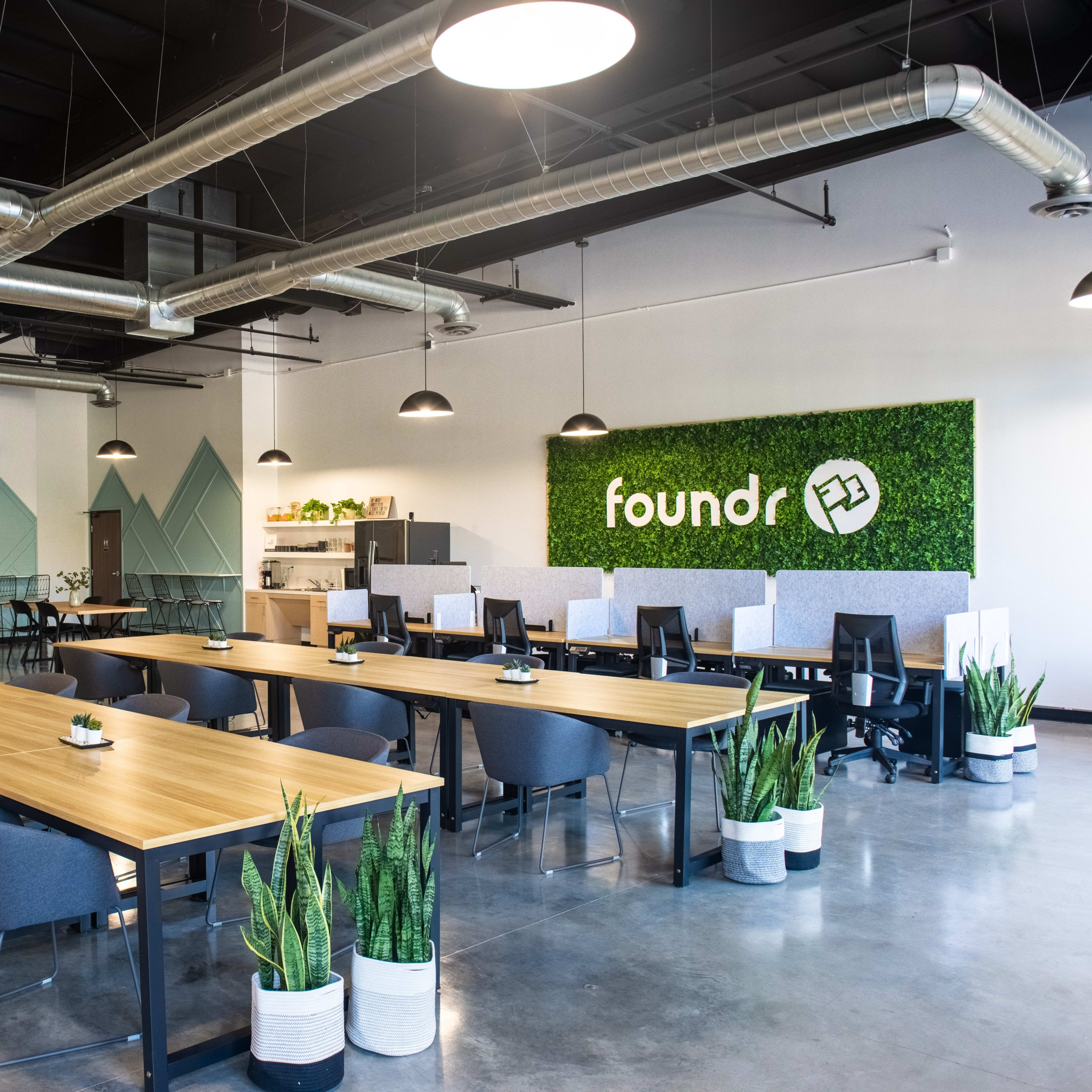 A modern office space with multiple workstations, large tables, and greenery decor on the walls.