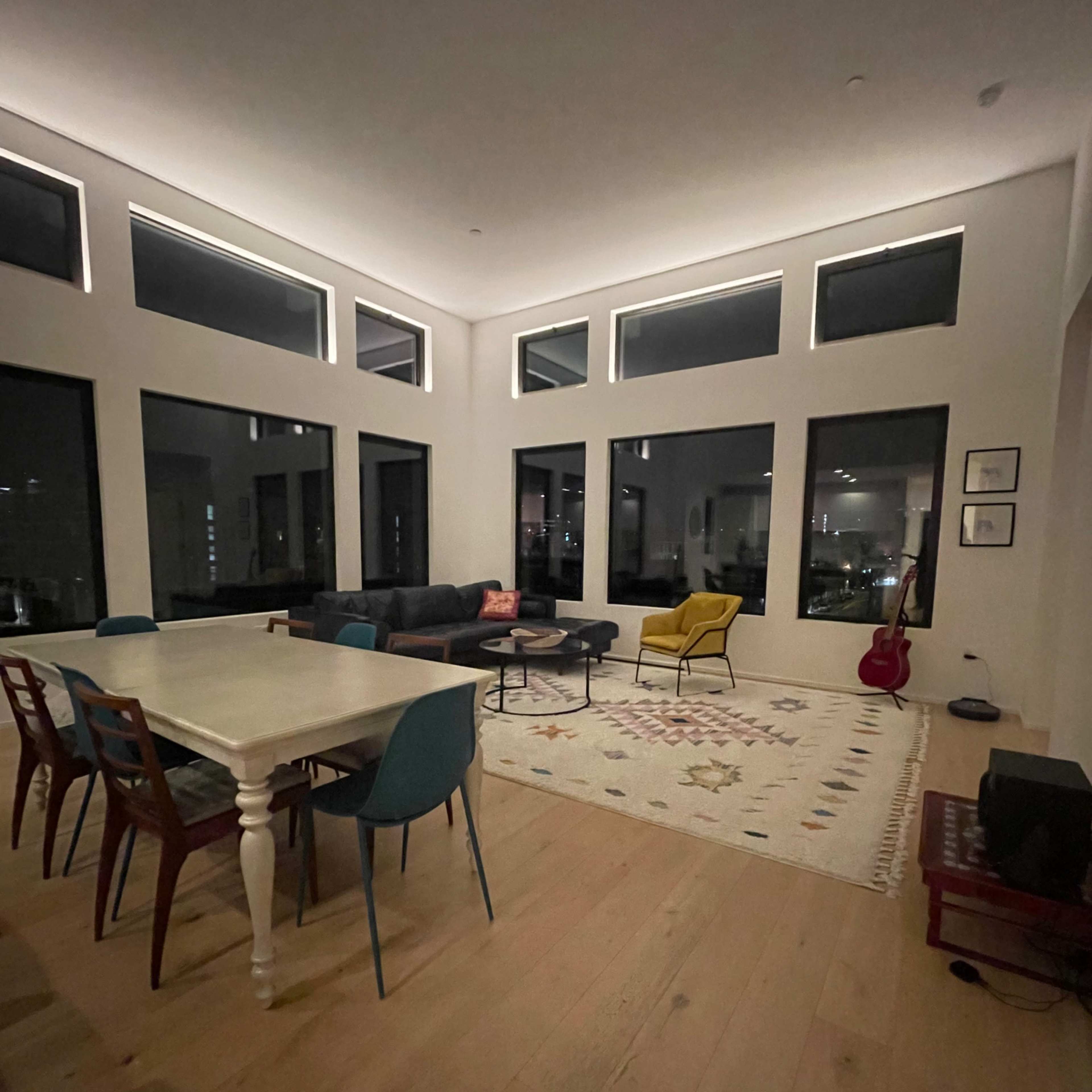 A modern living space features a dining table with colorful chairs, a dark sectional couch, a yellow armchair, and large windows allowing light to enter.