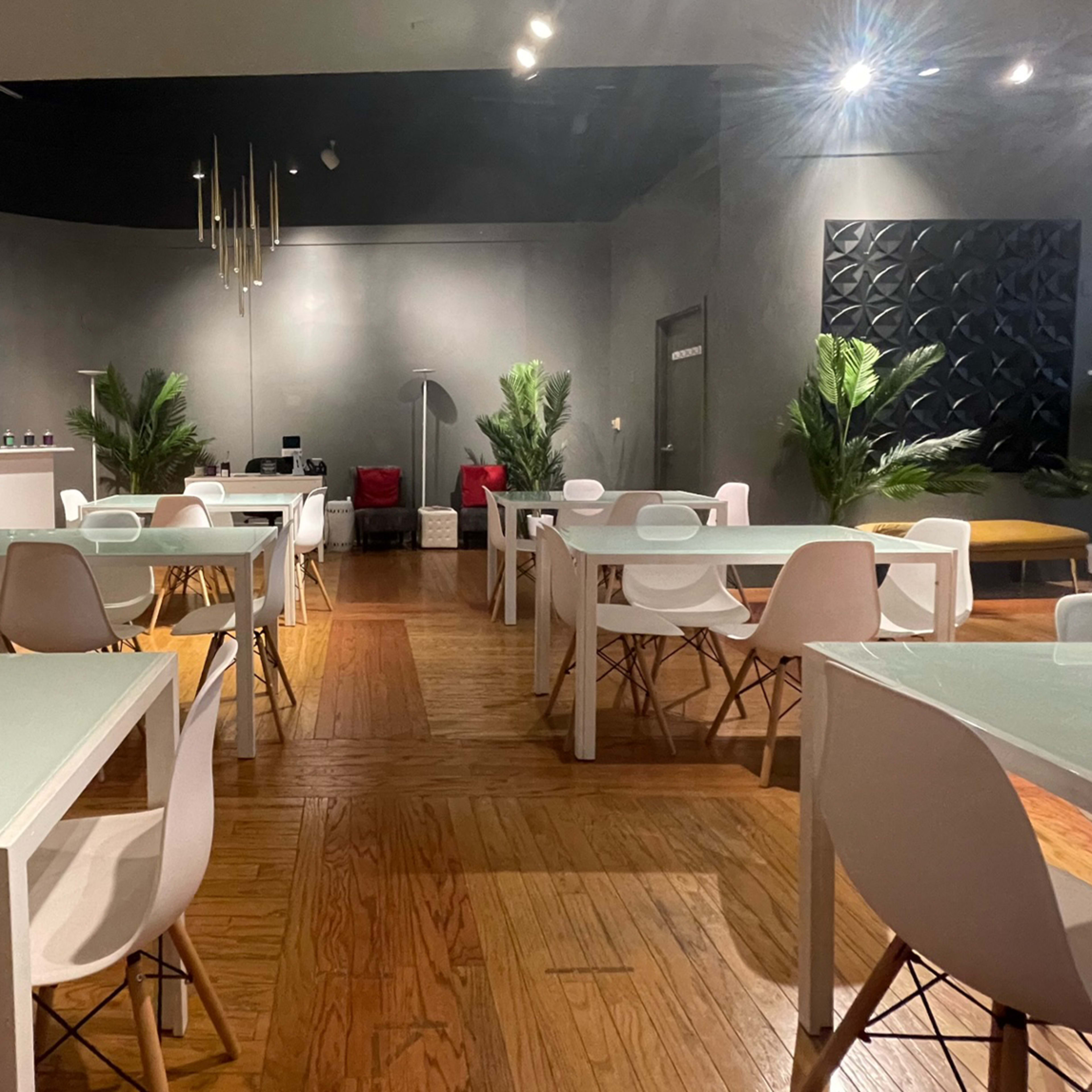 A modern, sparsely furnished interior with white tables and chairs, wooden flooring, and a few decorative plants.