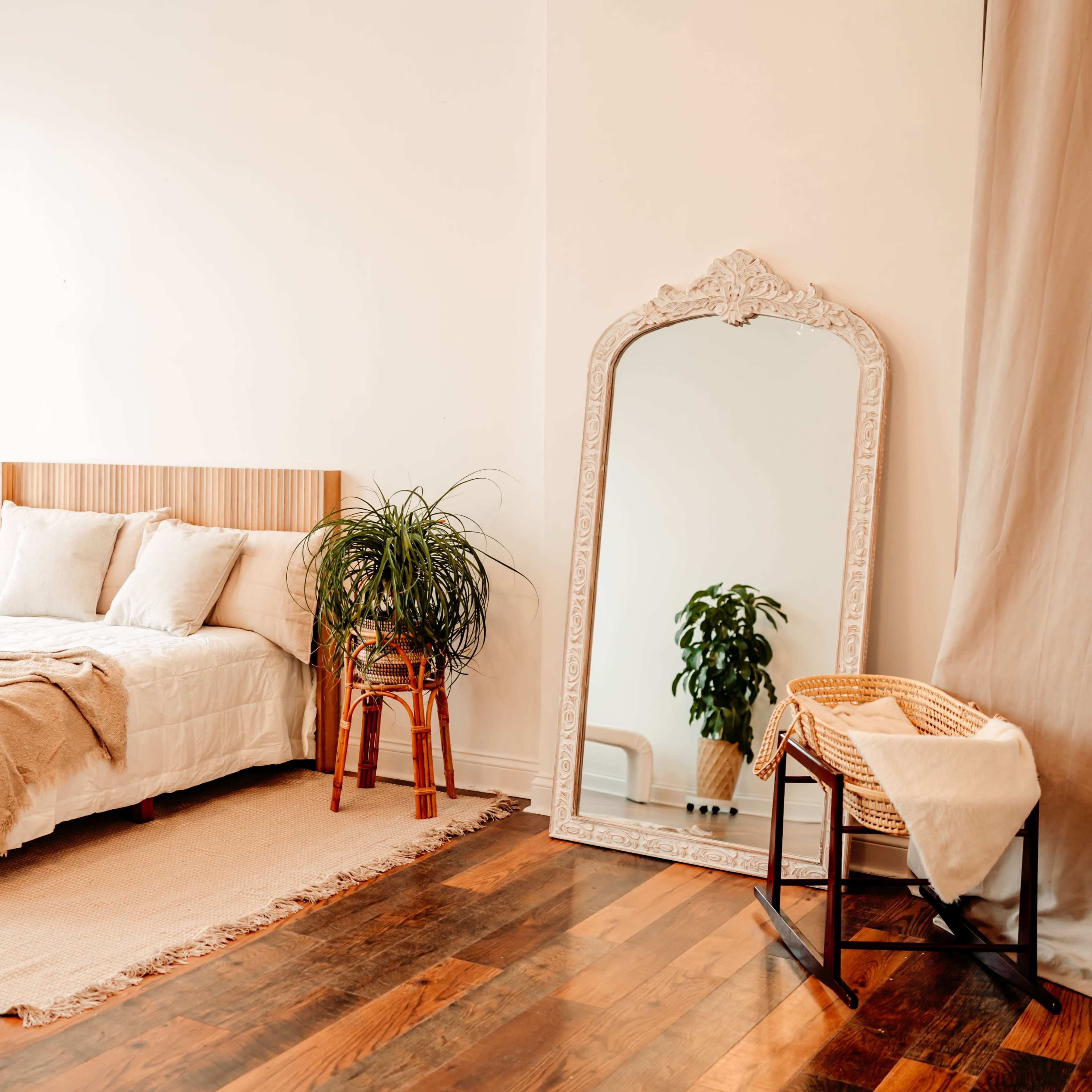 A cozy bedroom with a wooden bed, a large mirror, a plant on a stand, and a woven basket next to a light-colored rug on a hardwood floor.