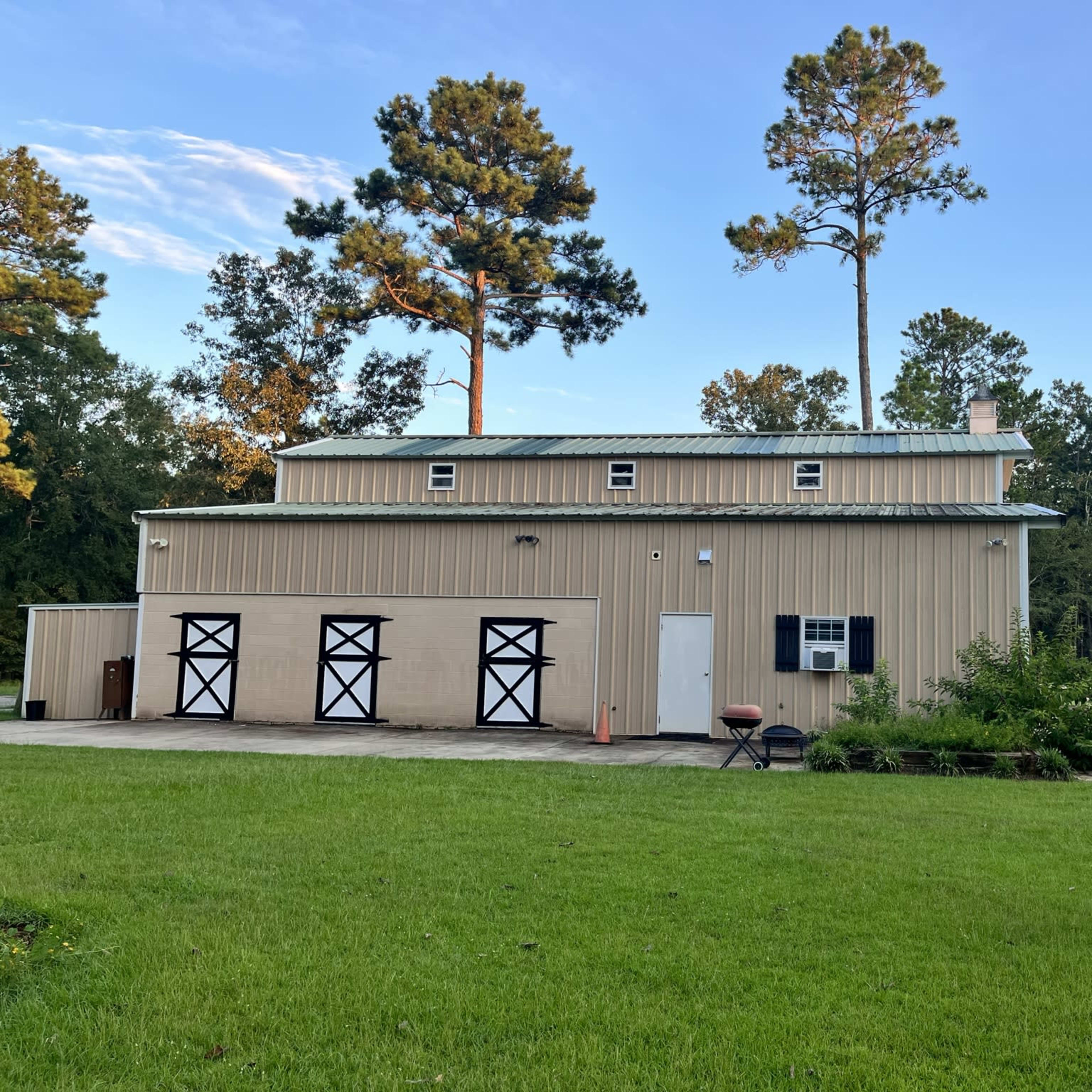 A large metal building with a tan exterior and three black garage doors is situated on a green lawn surrounded by trees.