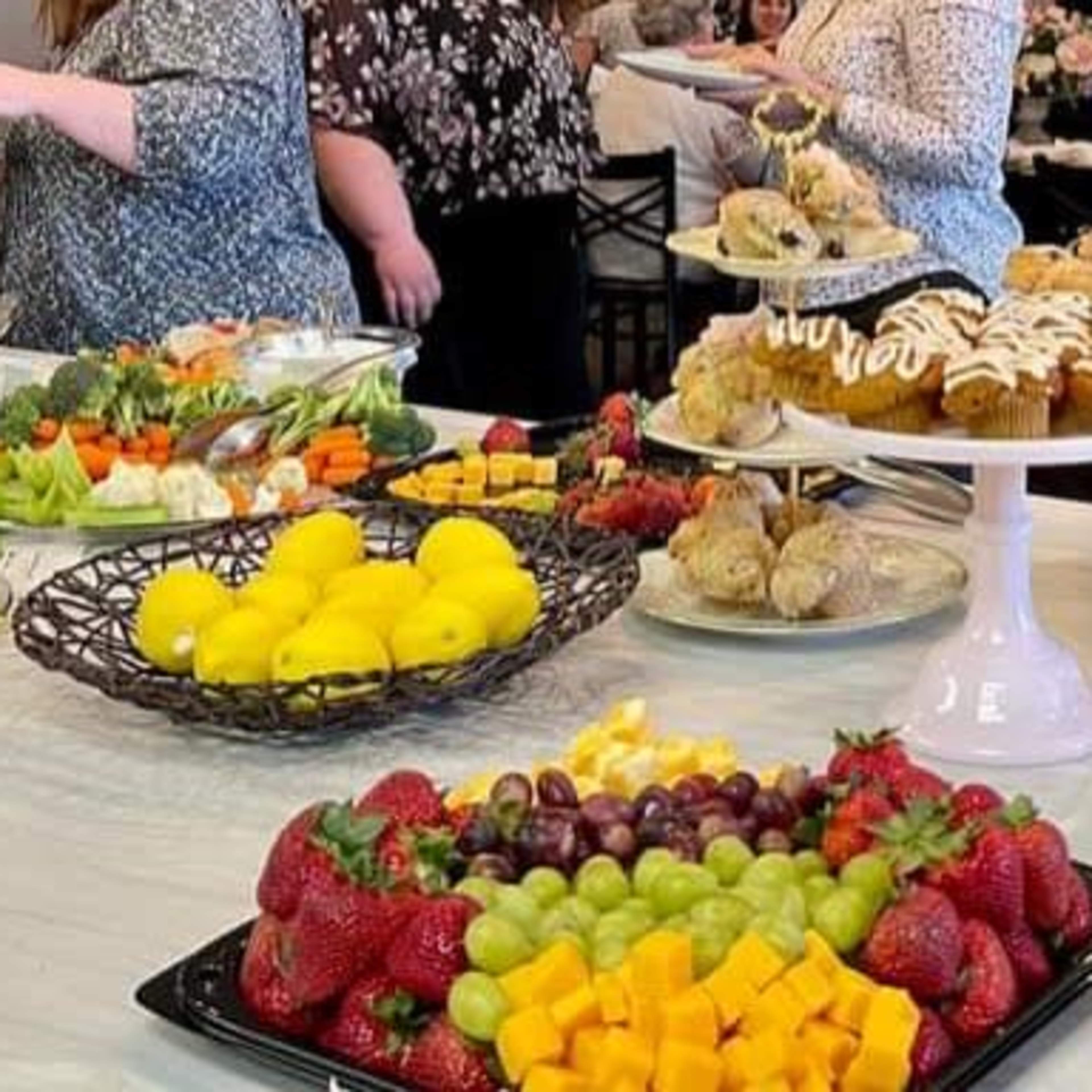 A buffet table is laden with an assortment of fruits, vegetables, baked goods, and desserts, while guests socialize in the background.