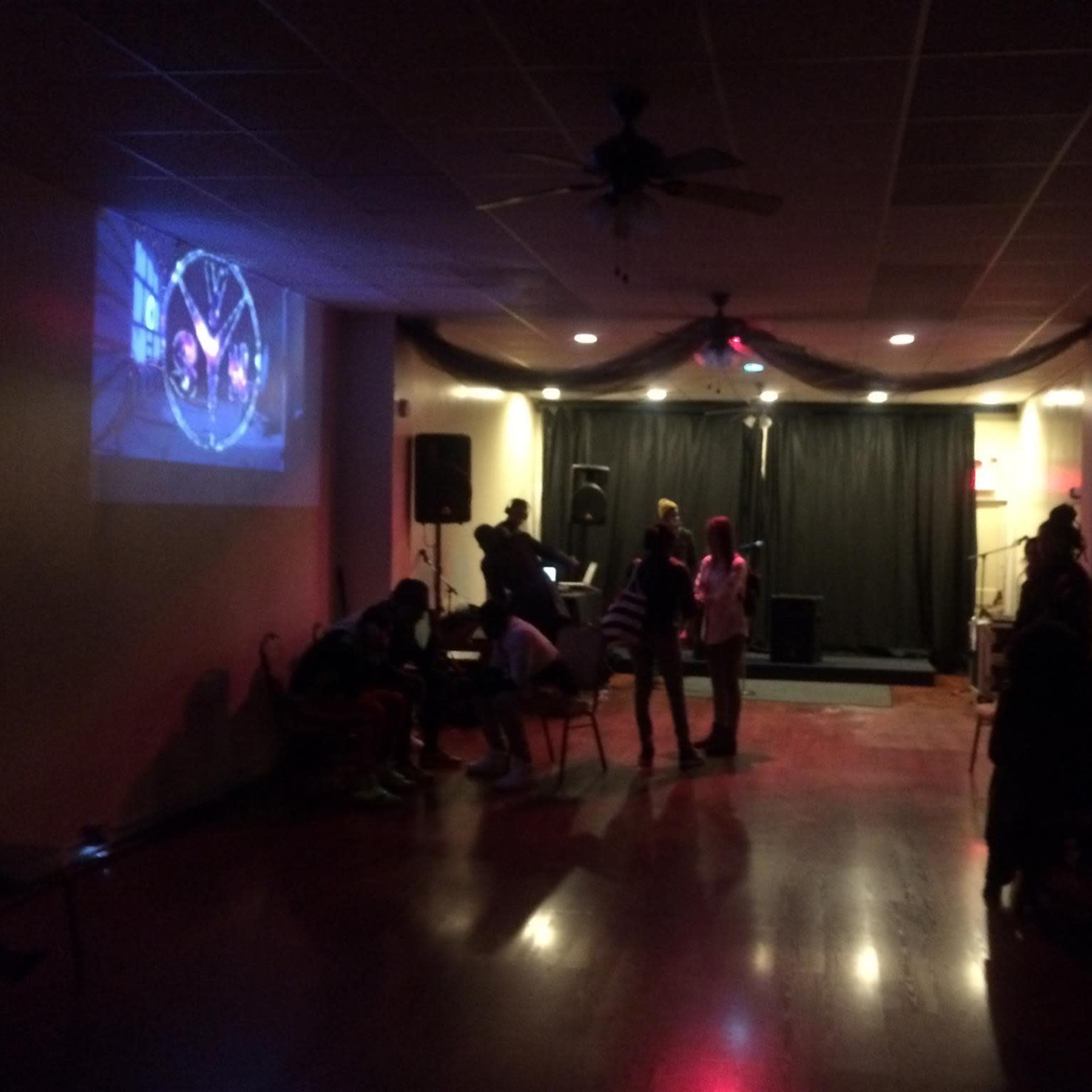 A gathering is taking place in a dimly lit room with a performance stage, chairs arranged around, and a video projected on a wall.