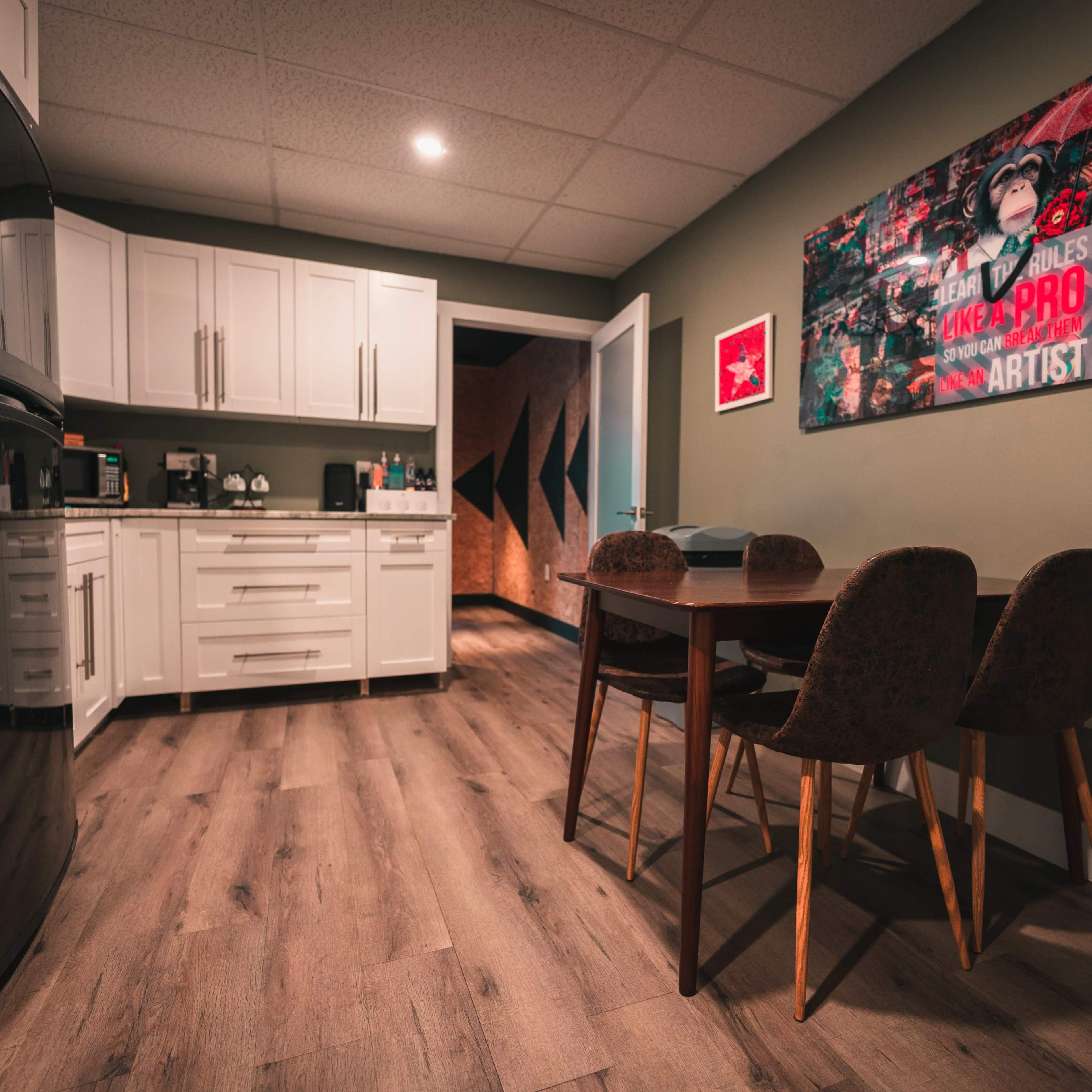 A modern kitchen area features white cabinets, a black refrigerator, a dining table with four chairs, and a vibrant artwork on the wall.