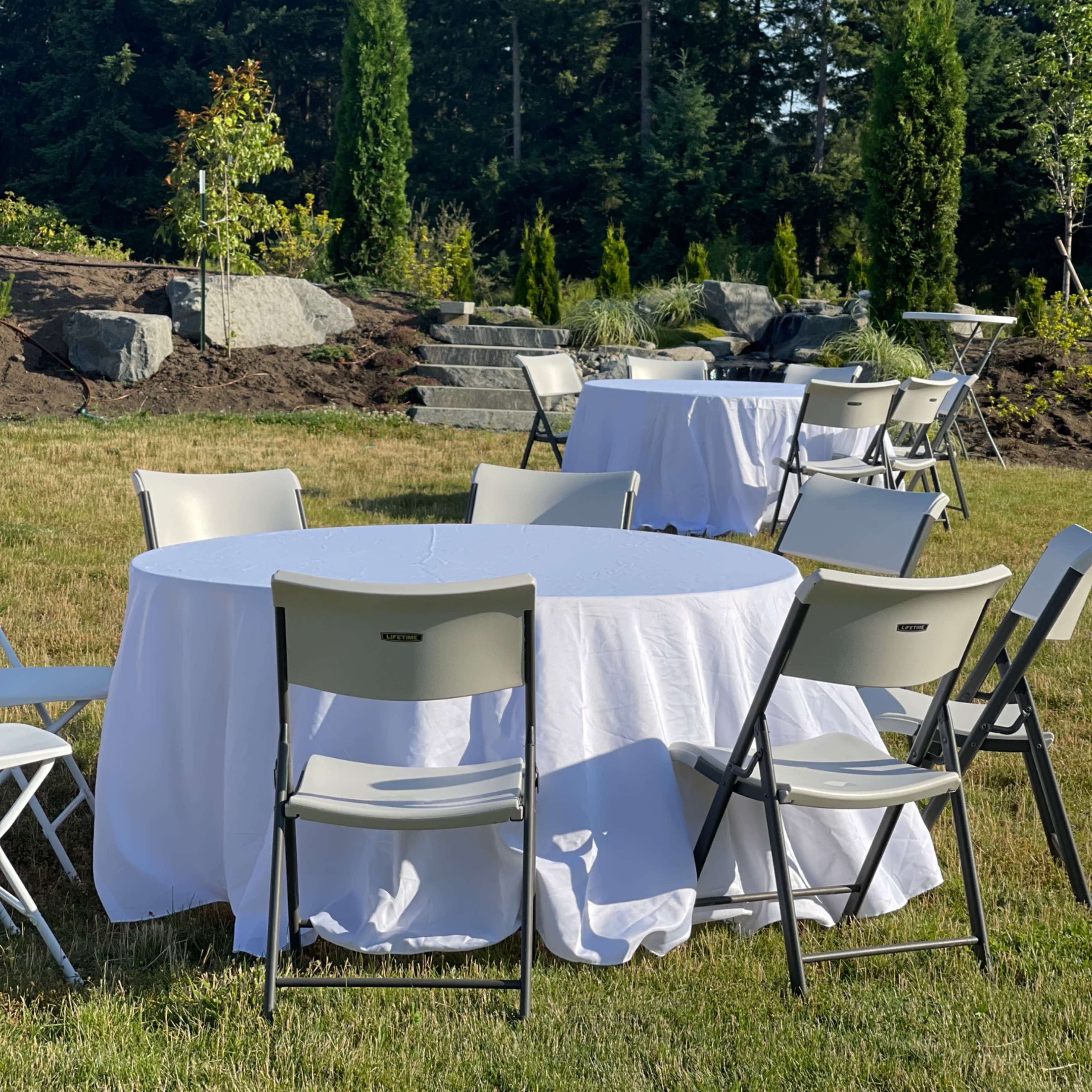 Tables covered with white tablecloths and surrounded by folding chairs are arranged on a grassy area with trees in the background.