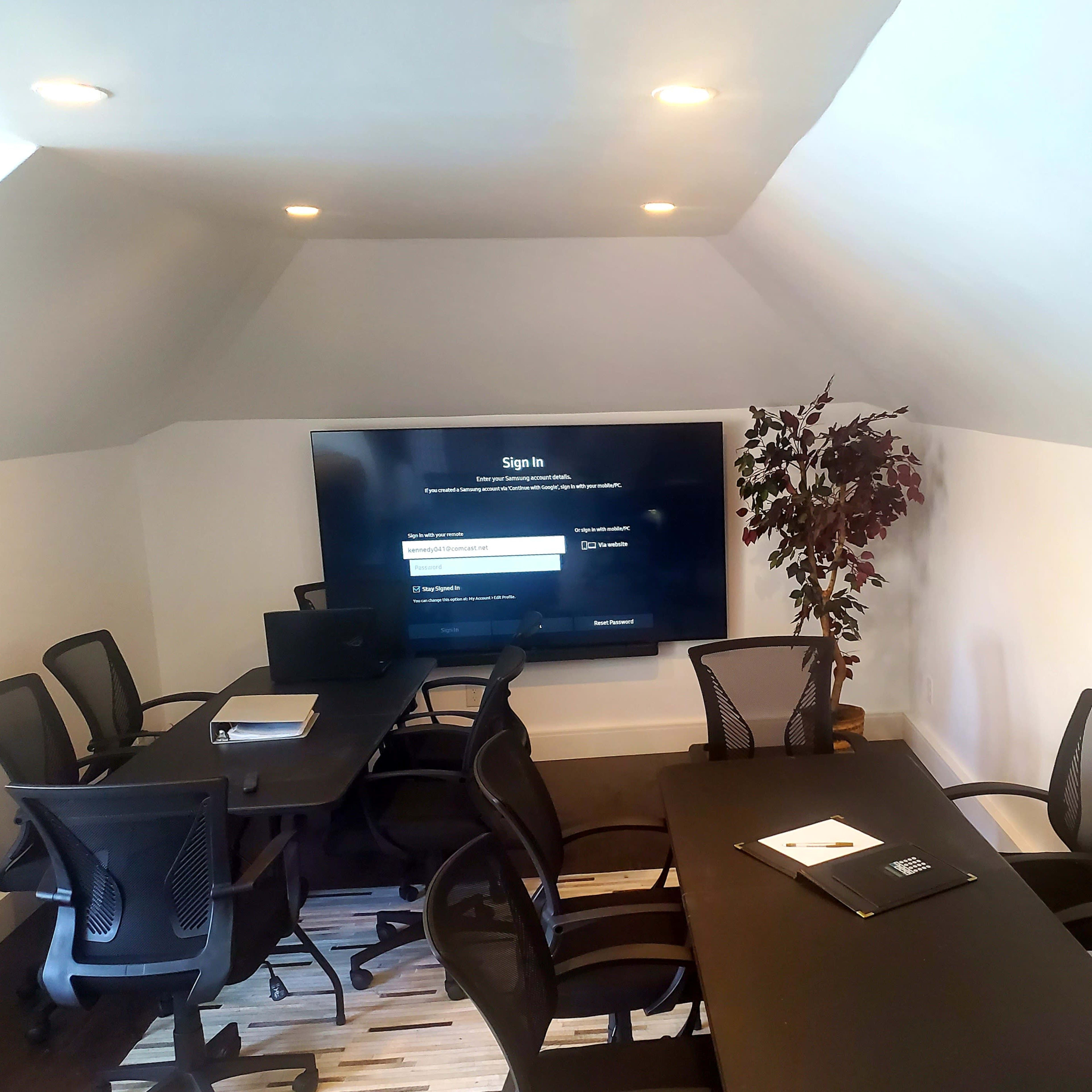 A meeting room features a large screen at one end, surrounded by several black chairs and a table, with a plant in the corner.