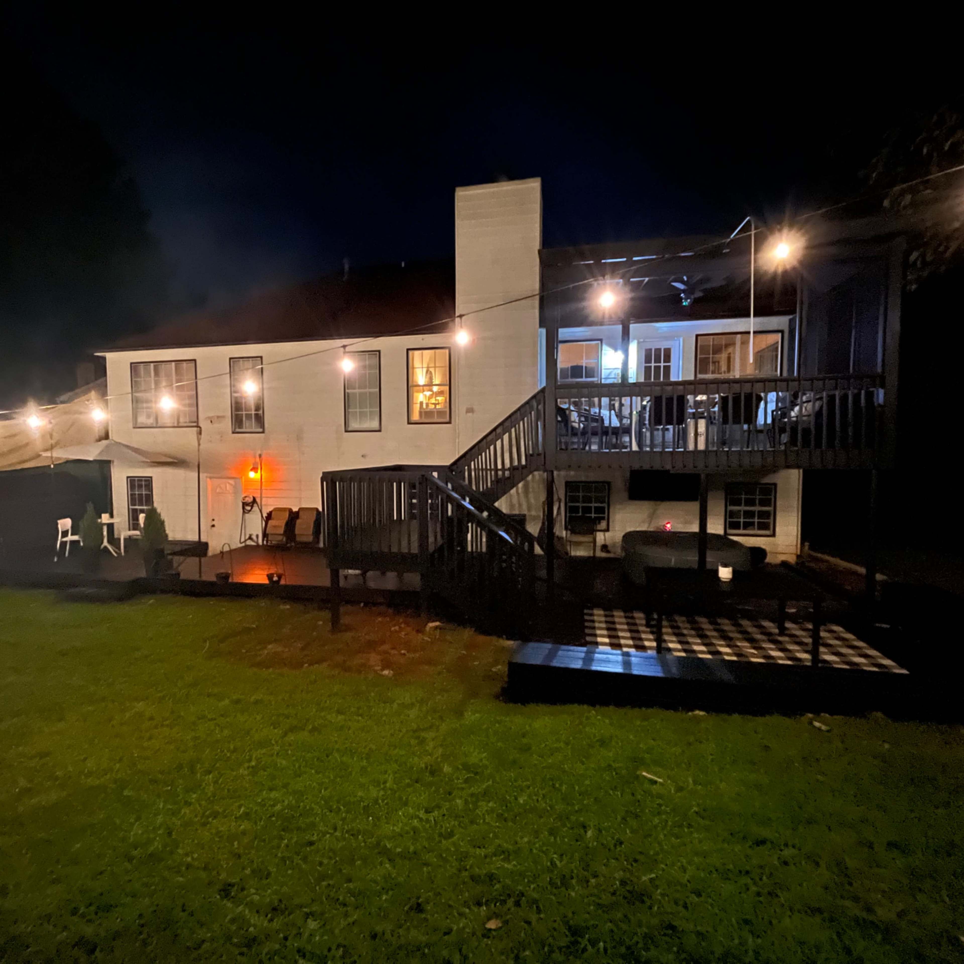 A two-story house with outdoor seating and string lights is illuminated at night, surrounded by a grassy yard.