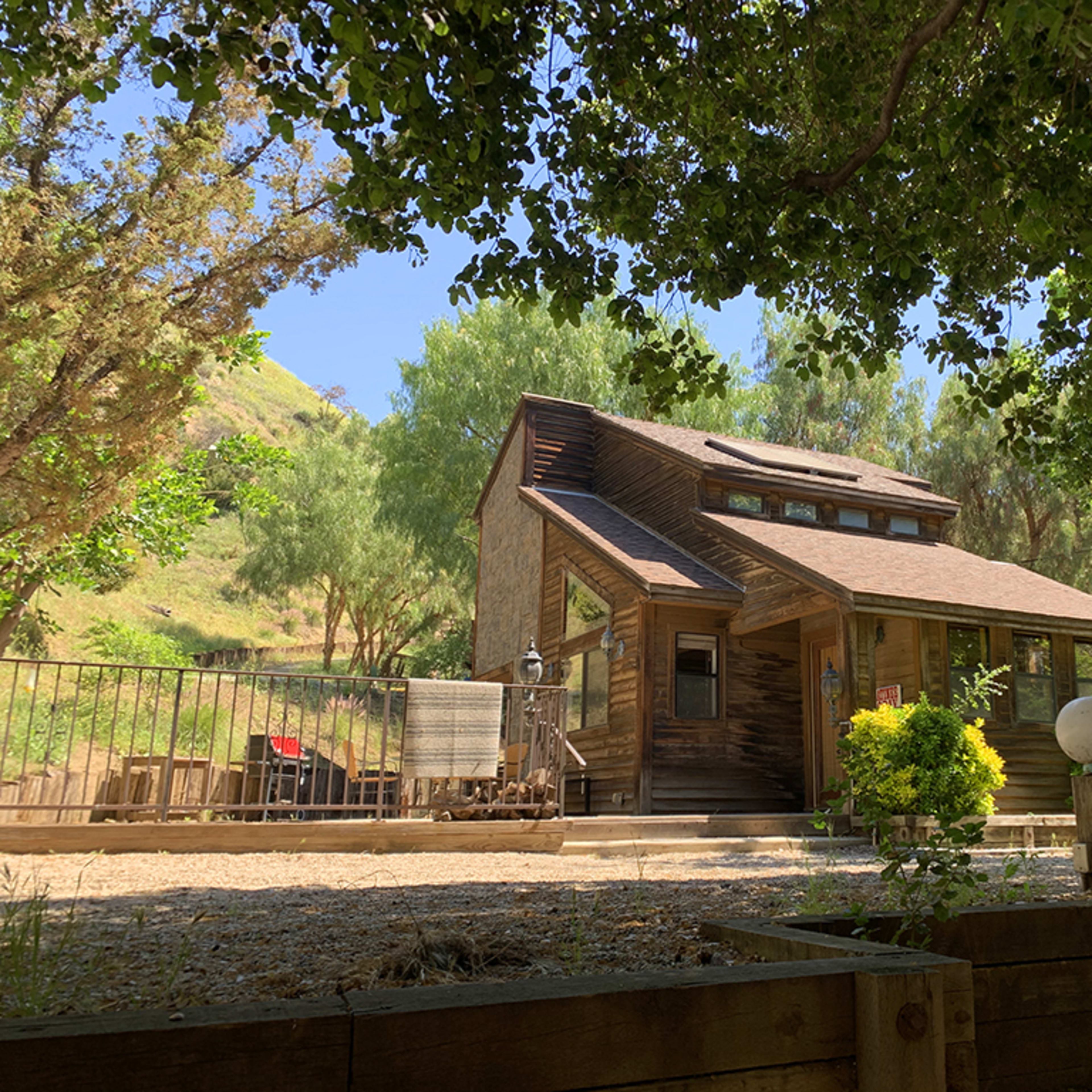 A wooden house with a sloped roof sits amidst trees on a hillside, featuring a fenced garden area in the foreground.