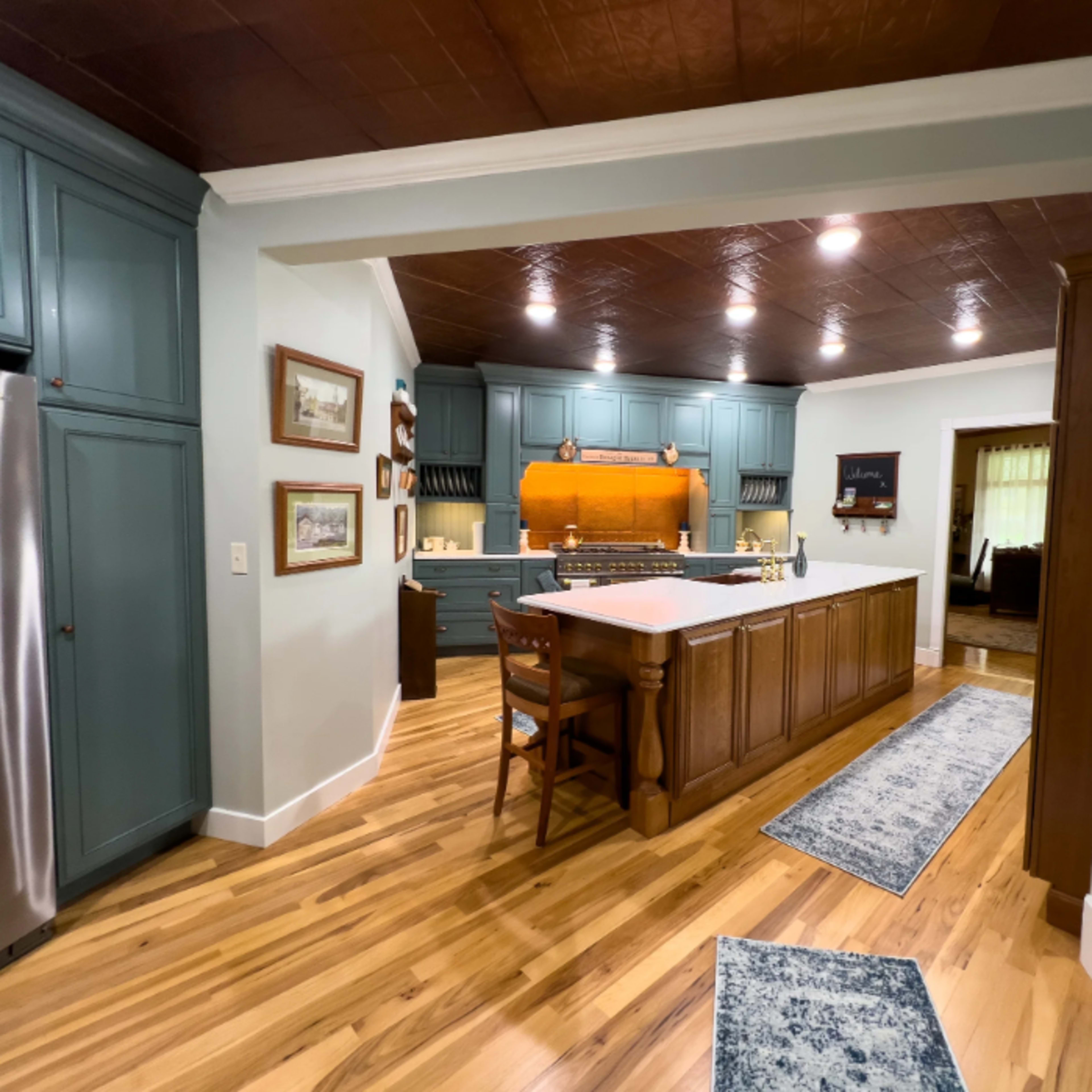 A spacious kitchen with teal cabinets, a central island with seating, hardwood flooring, and framed artwork on the walls.