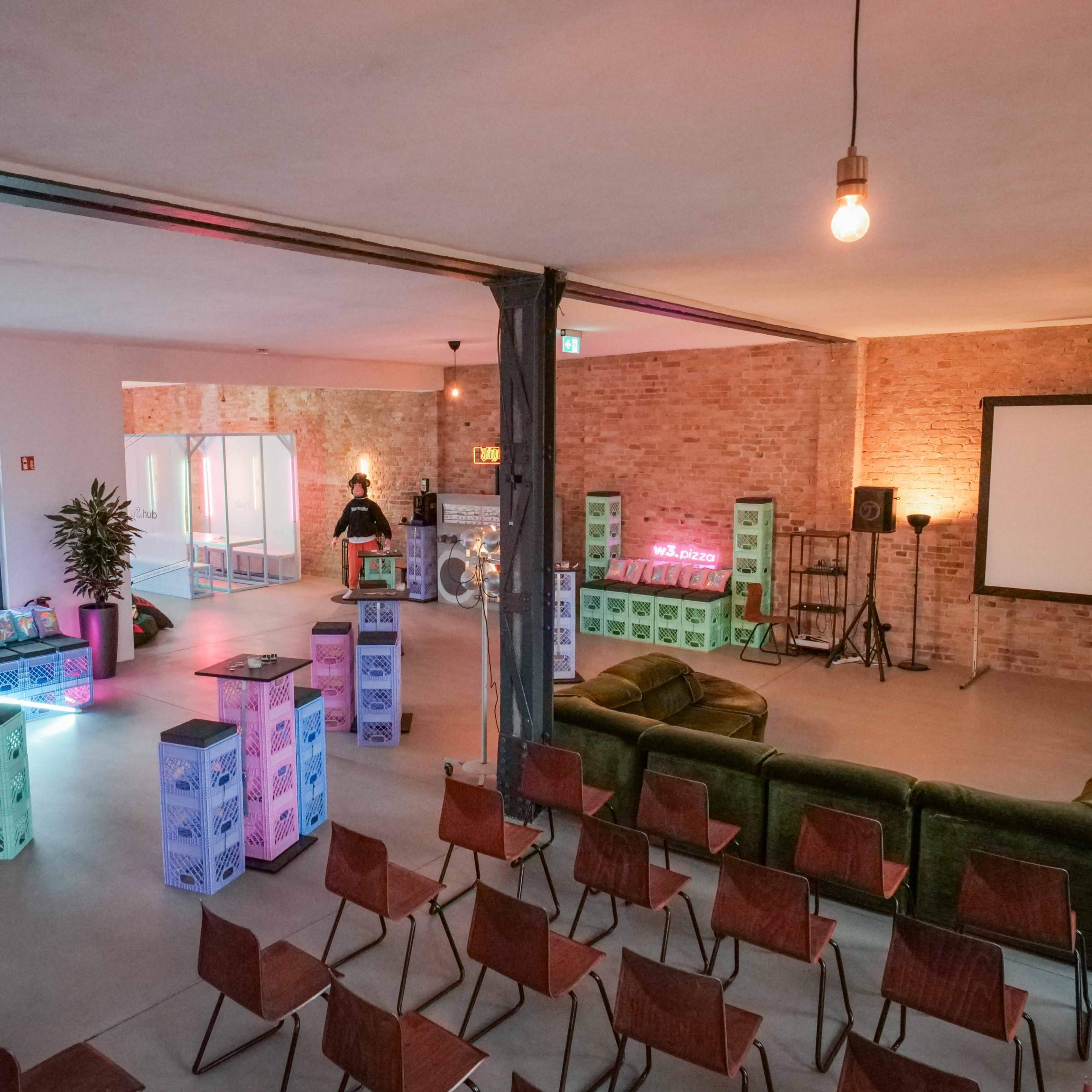 A spacious indoor venue features a mix of colorful seating areas, crates, and a projector screen, designed for social gatherings or events.