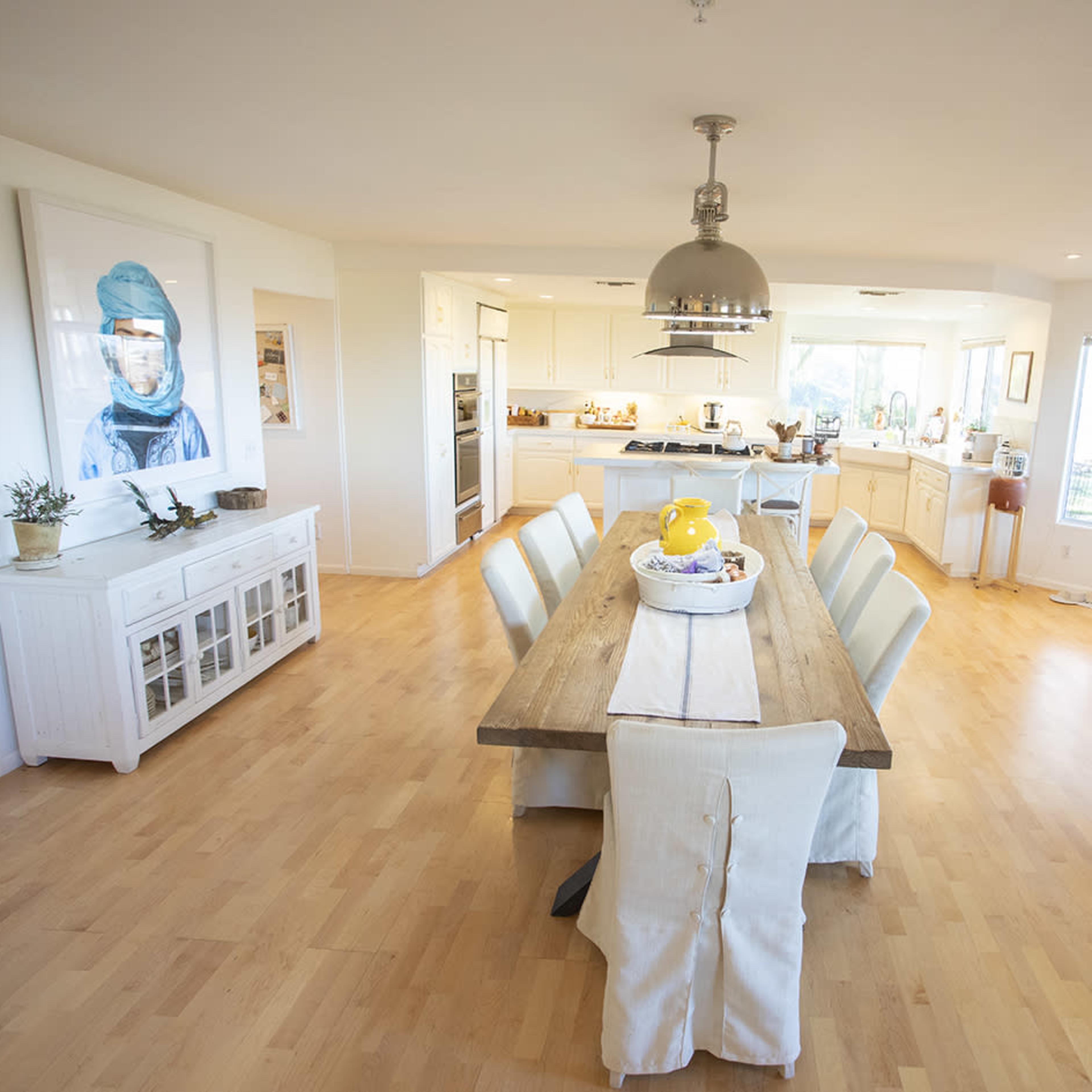 A spacious, light-filled dining area features a long wooden table surrounded by upholstered chairs, with a view of a kitchen in the background.