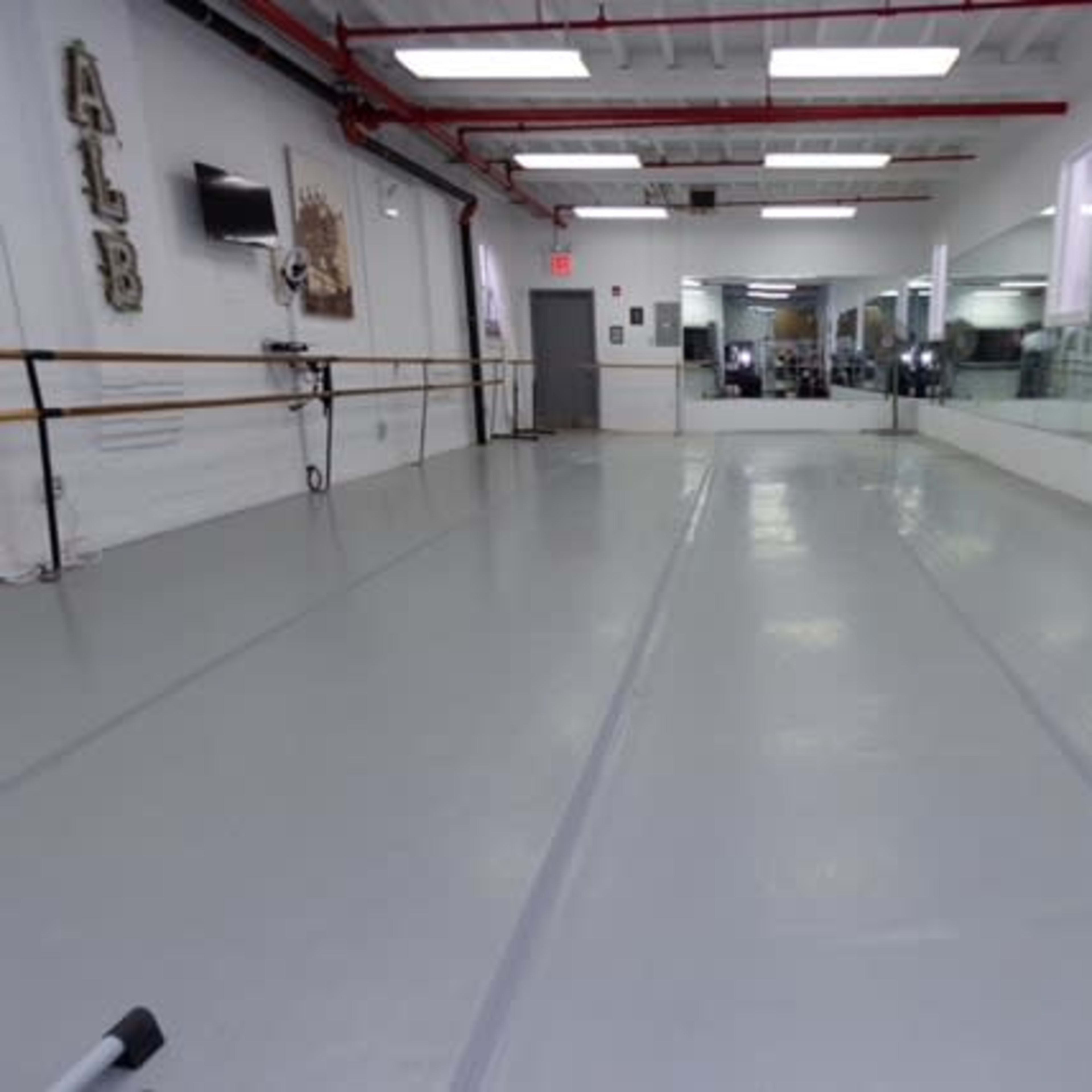 A spacious dance studio featuring a gray floor, mirrors along one wall, and ballet bars mounted on the opposite wall.