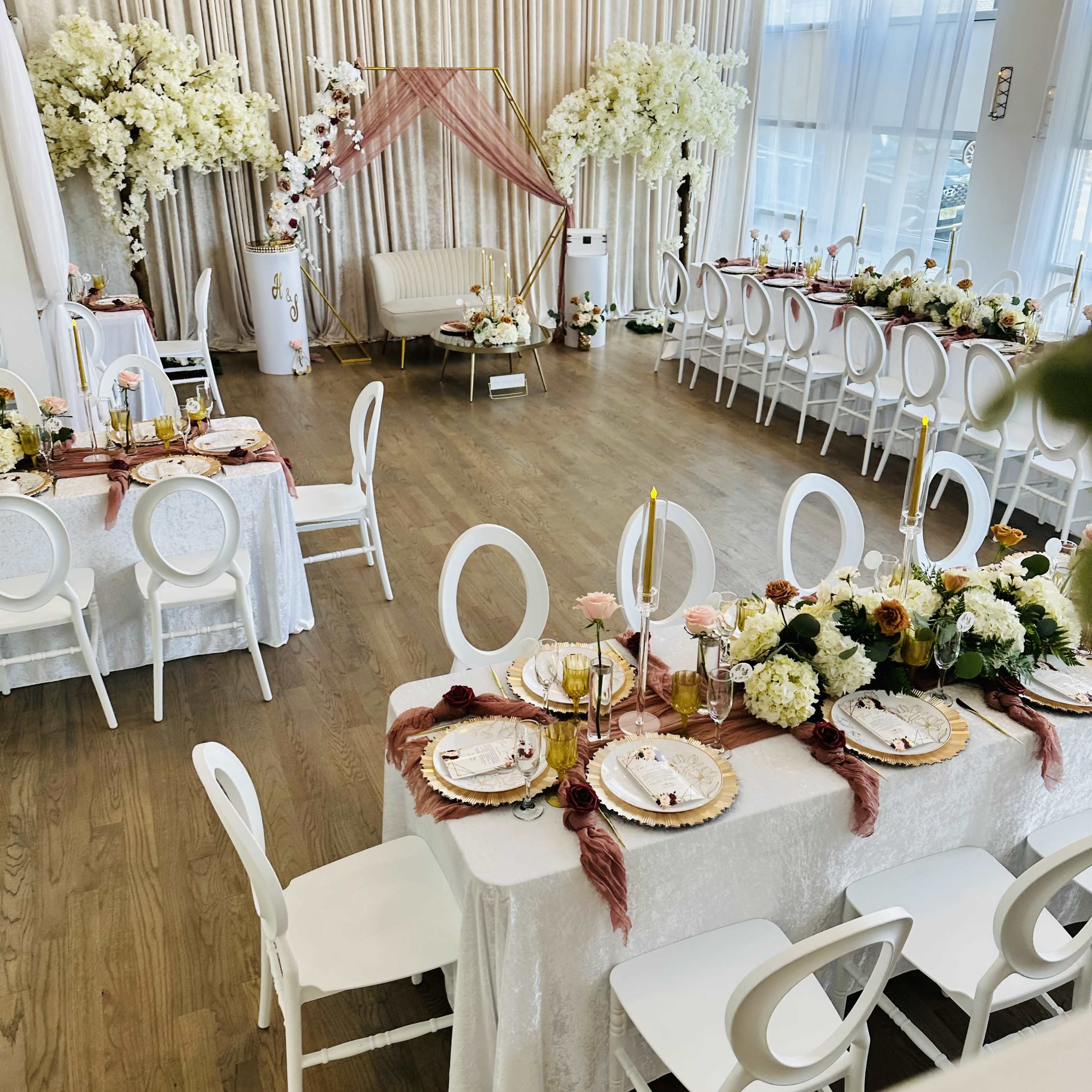 A elegantly arranged banquet hall featuring tables with white tablecloths, floral centerpieces, and decorative elements set for an event, with additional seating around the room.