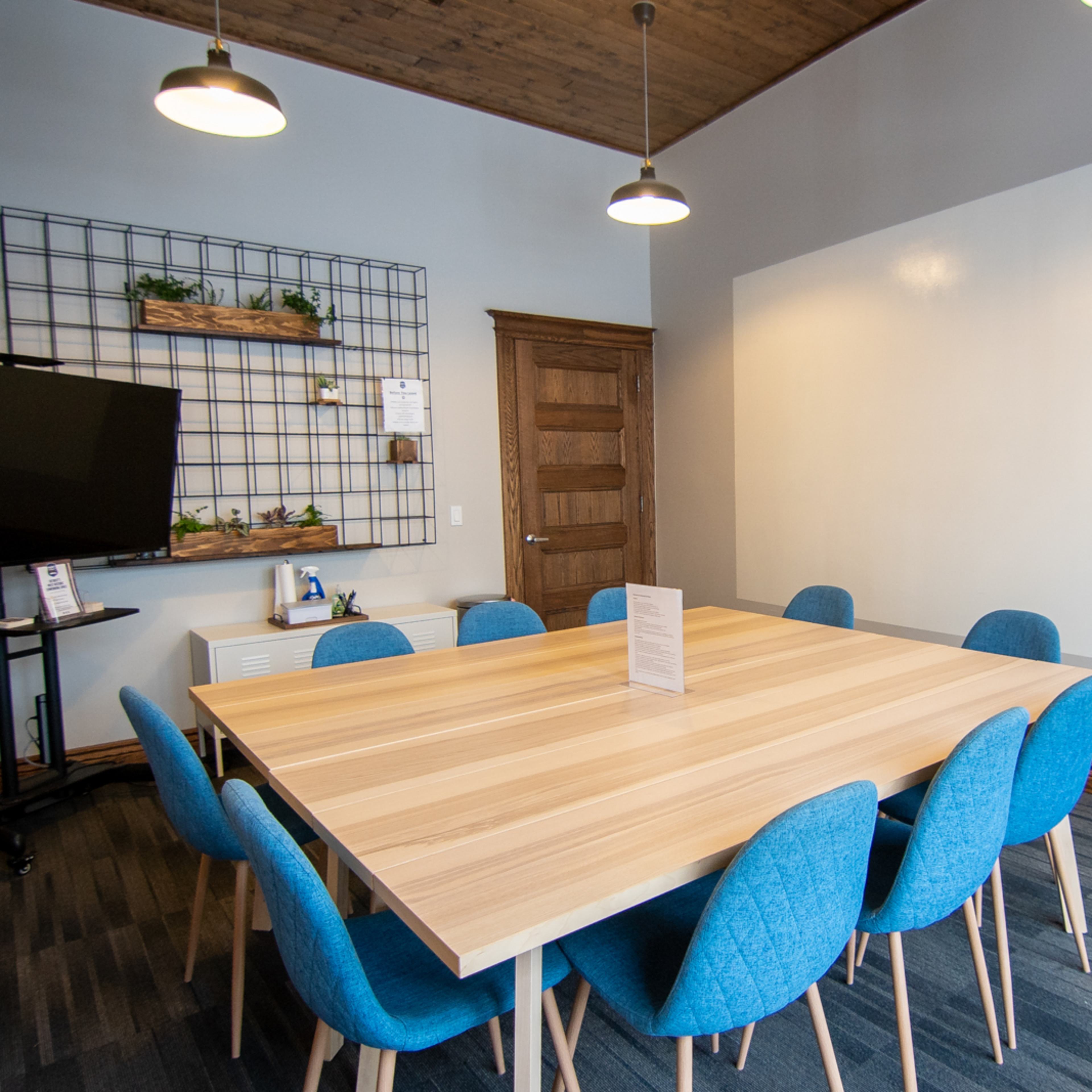 A meeting room features a large wooden table surrounded by eight blue chairs, with a wall-mounted shelf and a TV in the background.