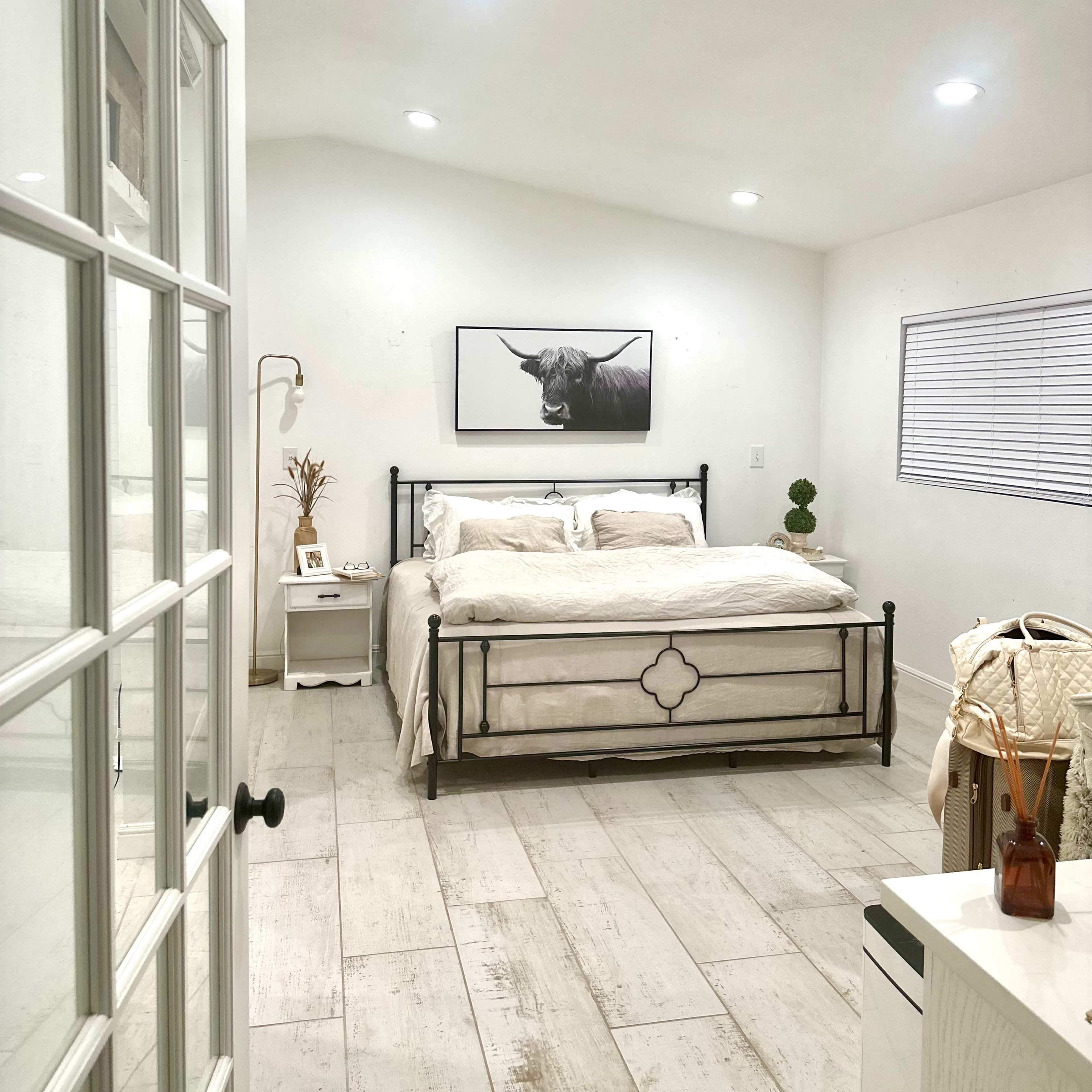 A bright bedroom features a metal bed frame with white bedding, a black-and-white bull print on the wall, and large windows with white blinds.