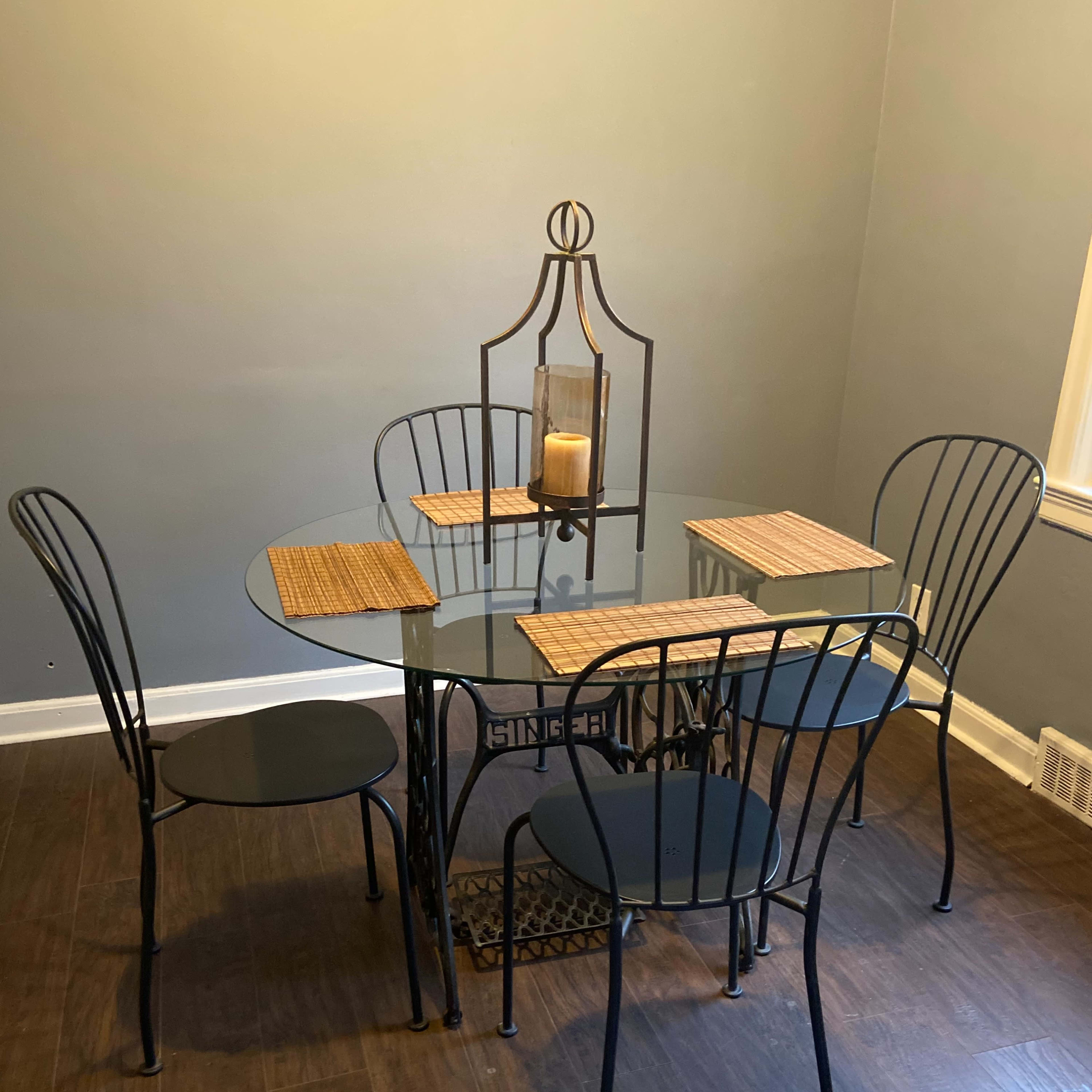 A glass-topped dining table is set with four black metal chairs and two placemats, with a candle in a lantern centerpiece.