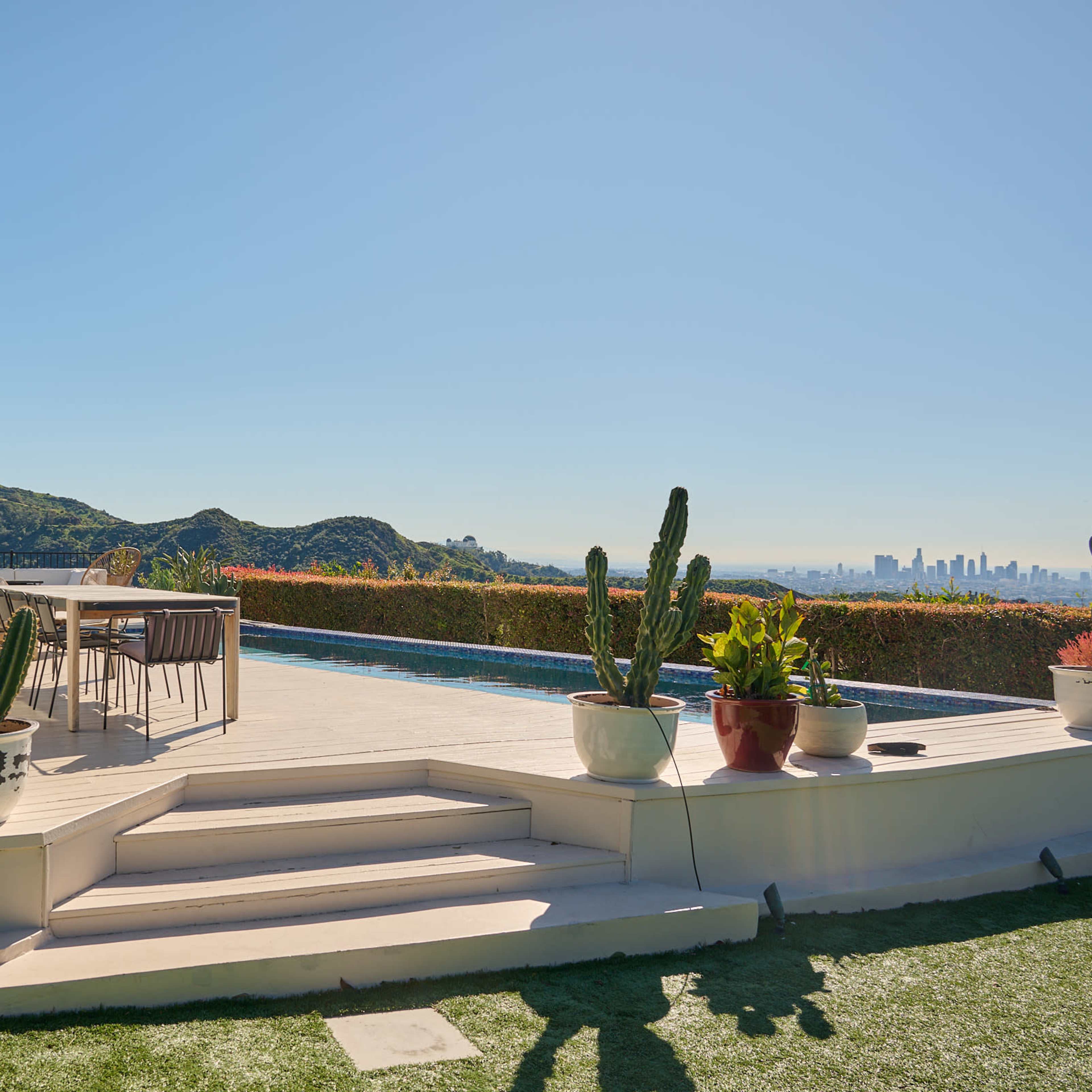 A spacious outdoor patio features potted plants and a swimming pool, set against a backdrop of hills and a distant city skyline.