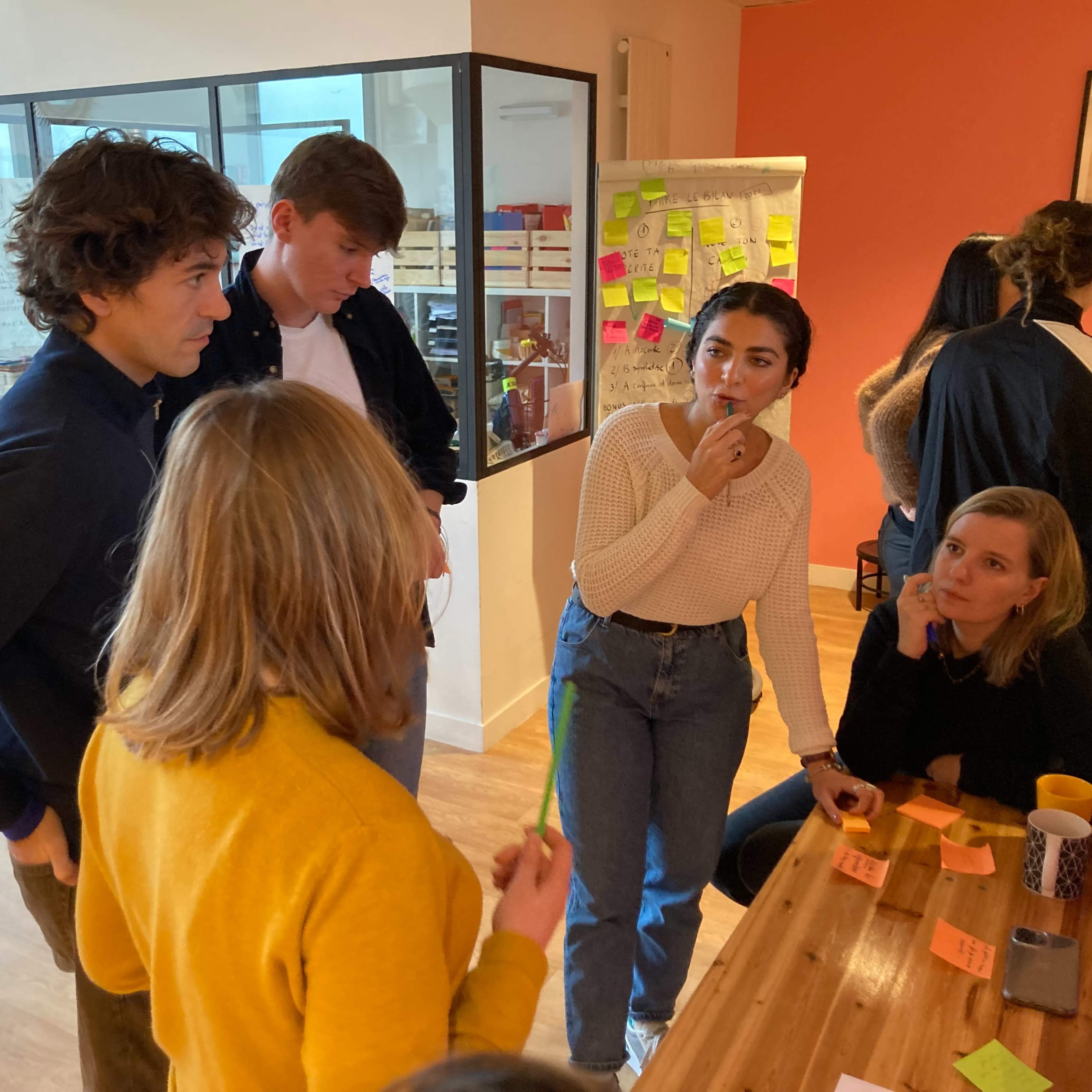A group of young adults engages in a discussion around a table filled with sticky notes in a brightly lit workspace.
