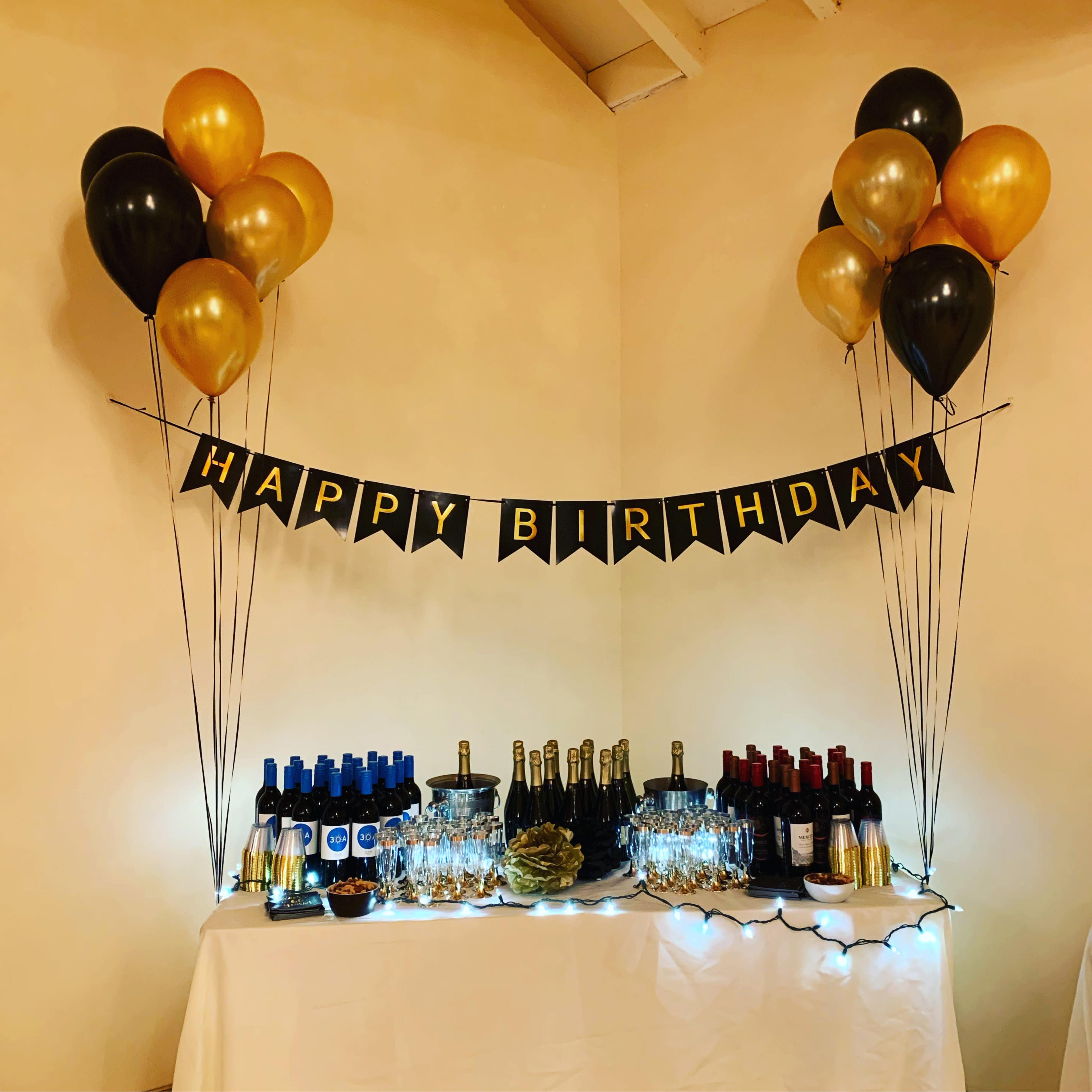 A birthday celebration setup featuring a table with various bottles of wine, champagne, glasses, and a banner that reads "HAPPY BIRTHDAY," surrounded by black, gold, and silver balloons.