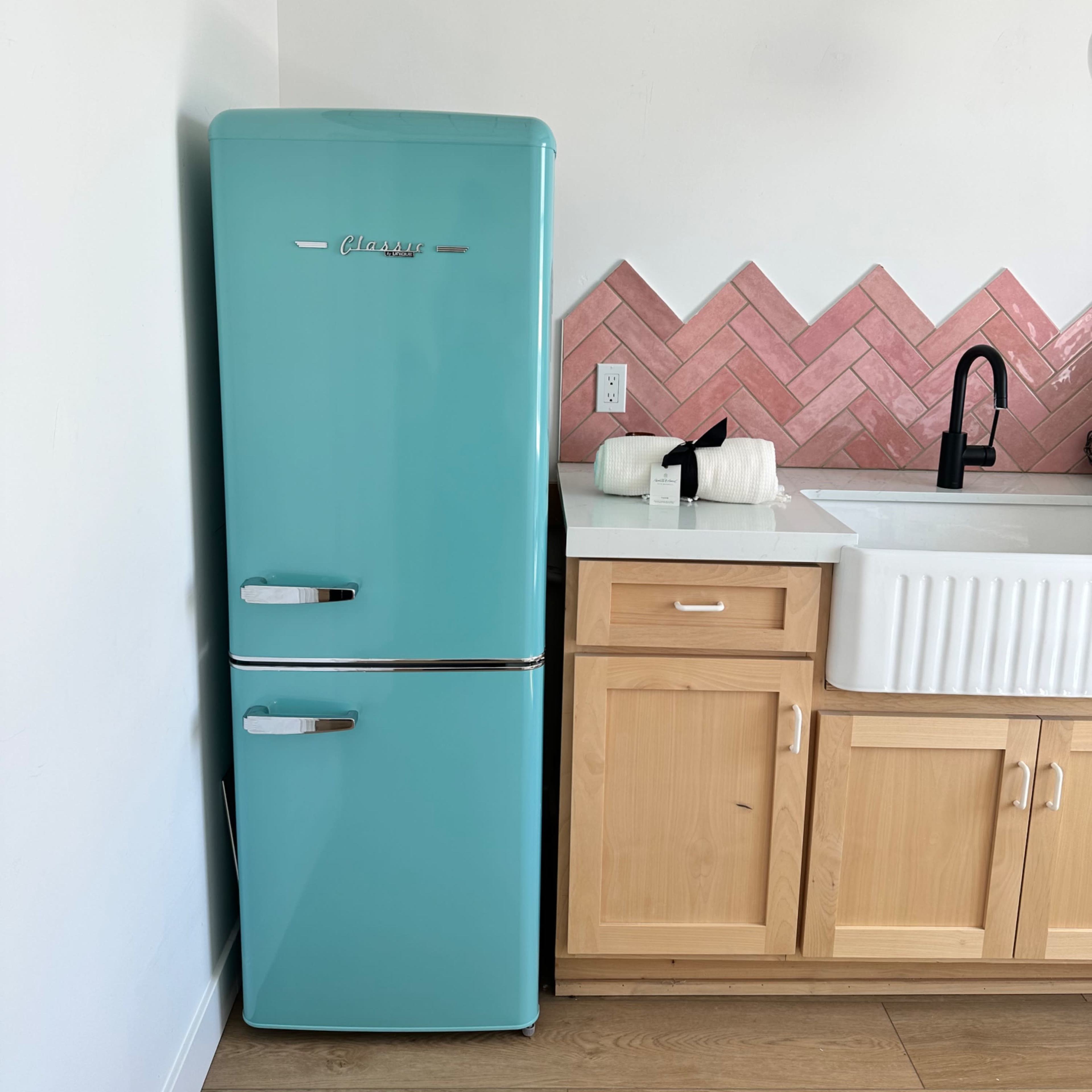 A teal retro refrigerator stands beside a white farmhouse sink and wooden cabinetry with pink herringbone tile backsplash.