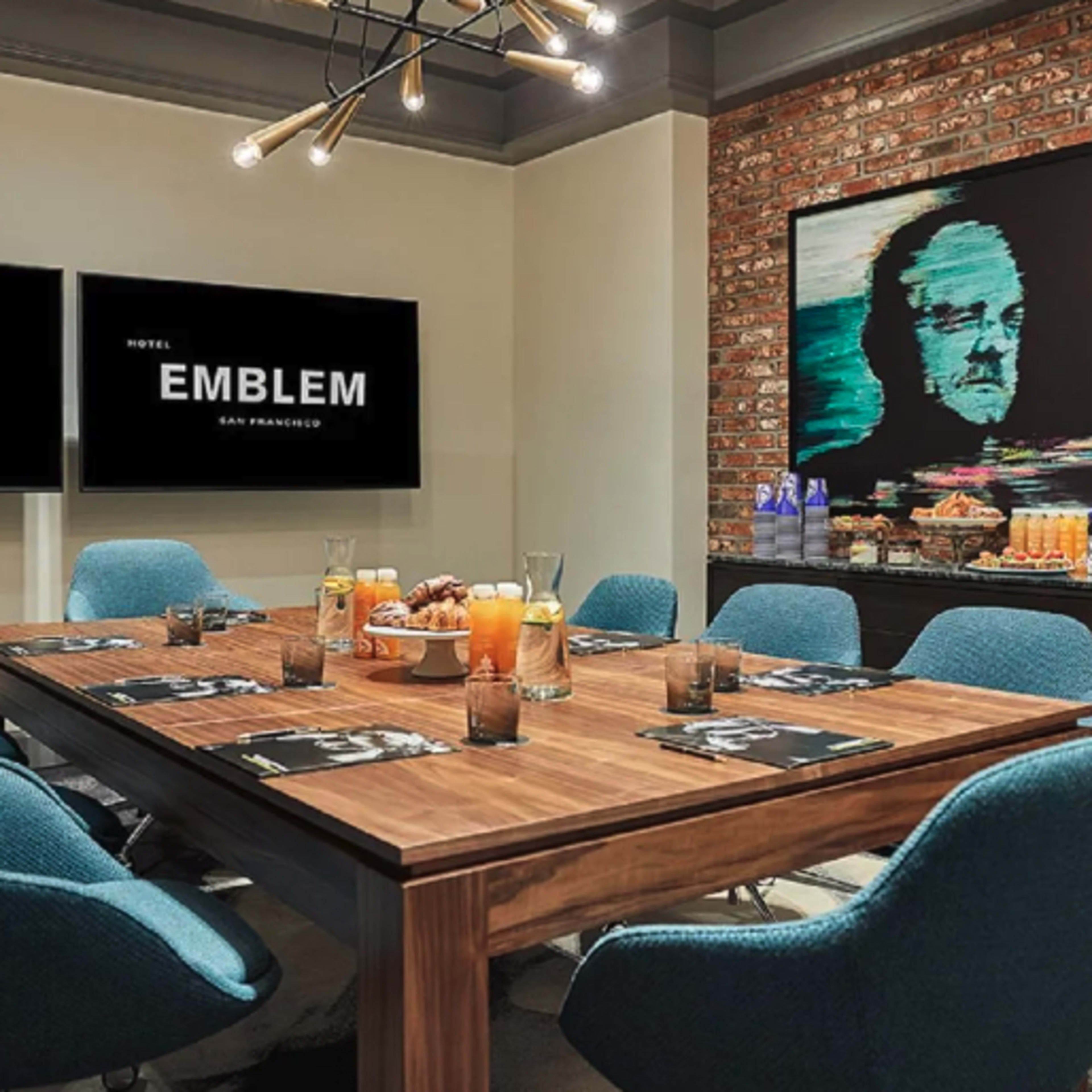 A conference room features a wooden table surrounded by teal chairs, with two screens displaying the hotel name and a large painting on the wall.