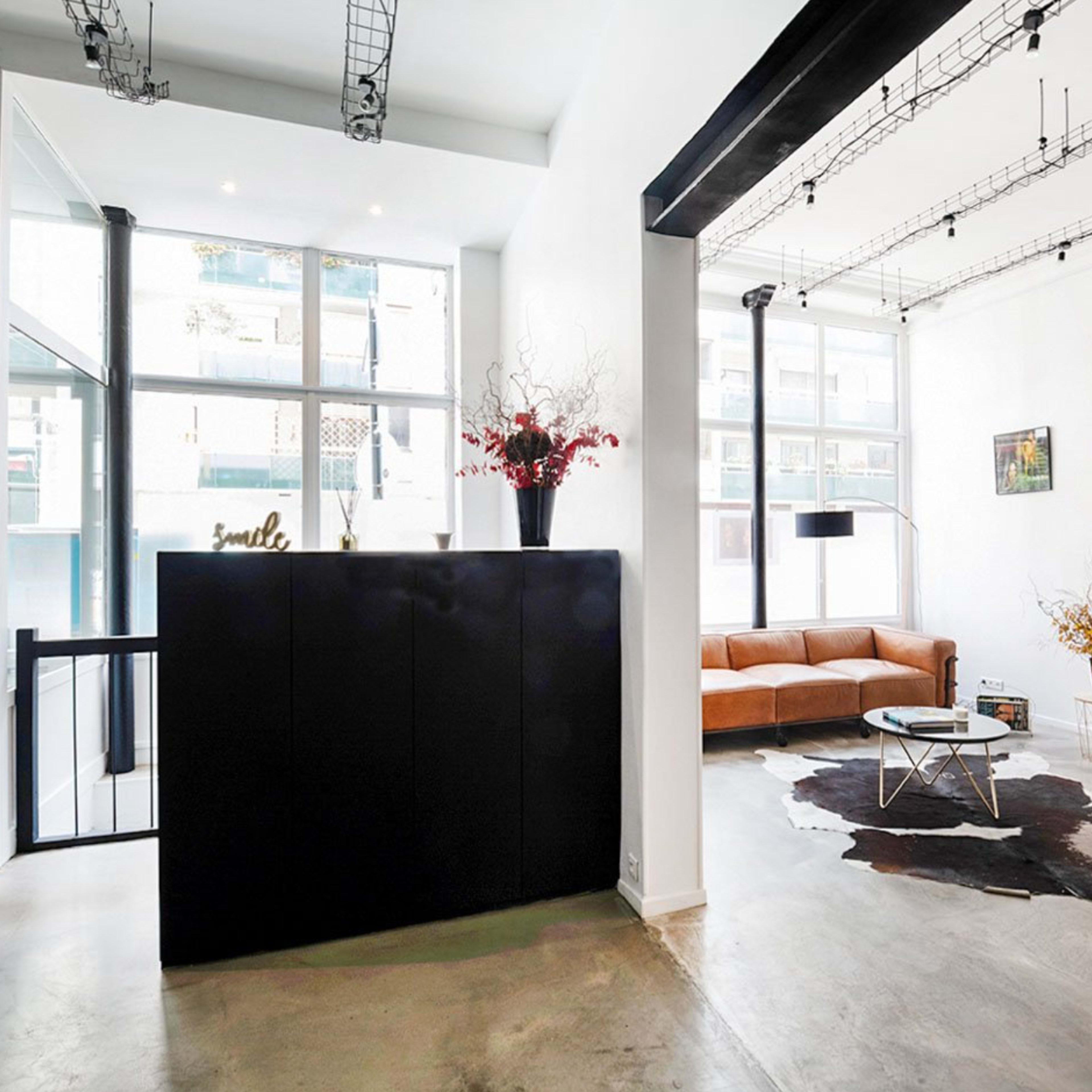 A modern interior space featuring a reception area with a black counter, a brown sofa, a coffee table on a cowhide rug, and large windows letting in natural light.