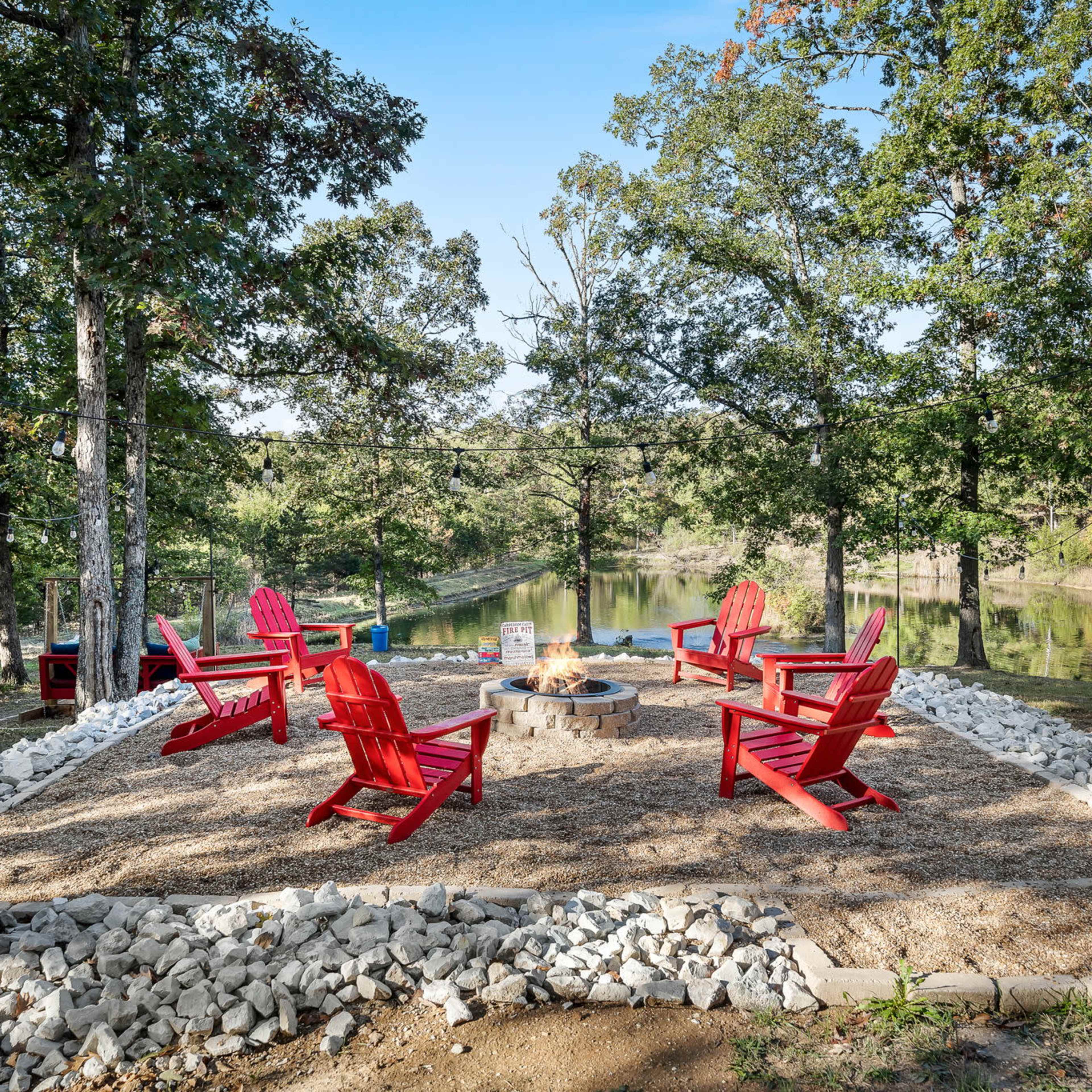 A circular seating arrangement of red Adirondack chairs surrounds a fire pit on a gravel and stone-lined area, set in a wooded location near a calm body of water.